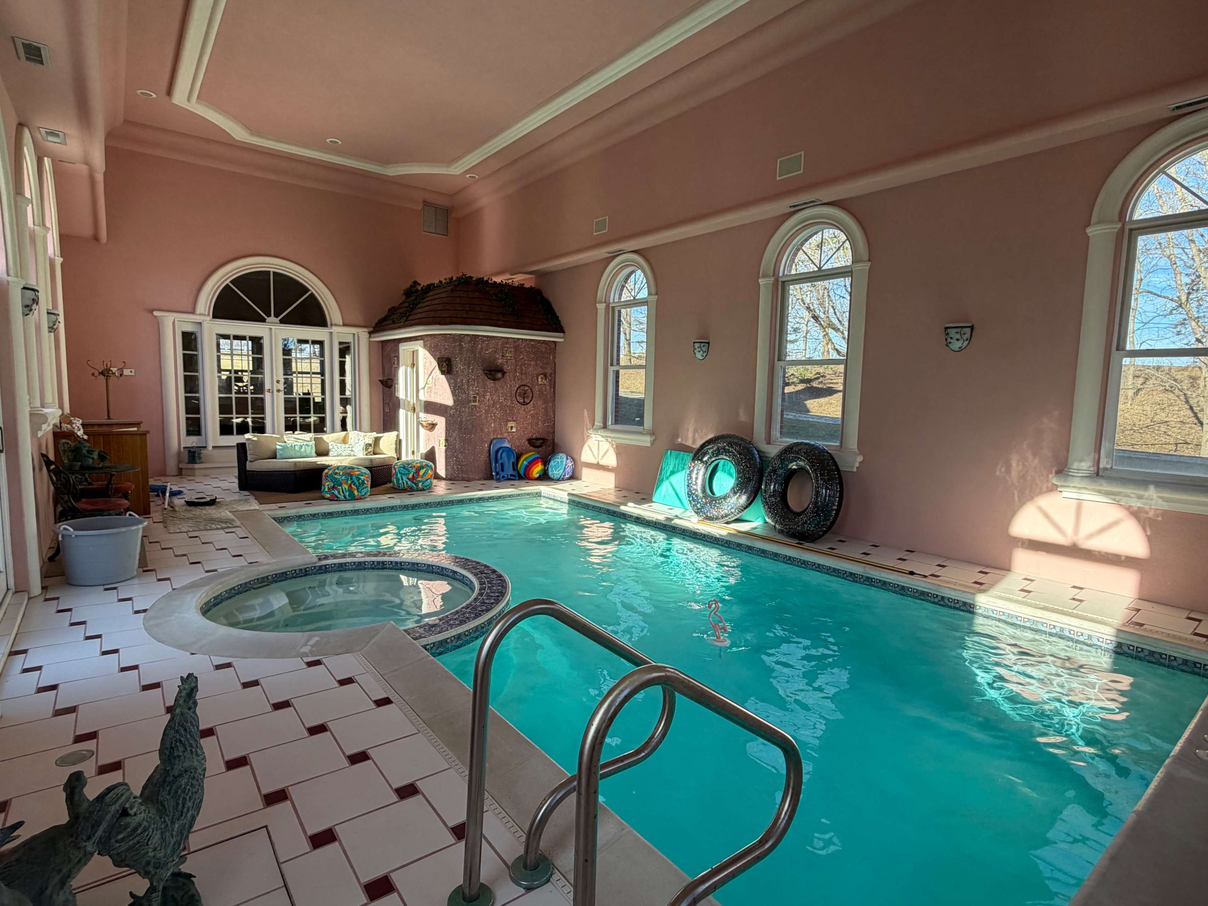 The image shows an indoor swimming pool area with a small spa, pink walls, and large arched windows allowing natural light to enter.