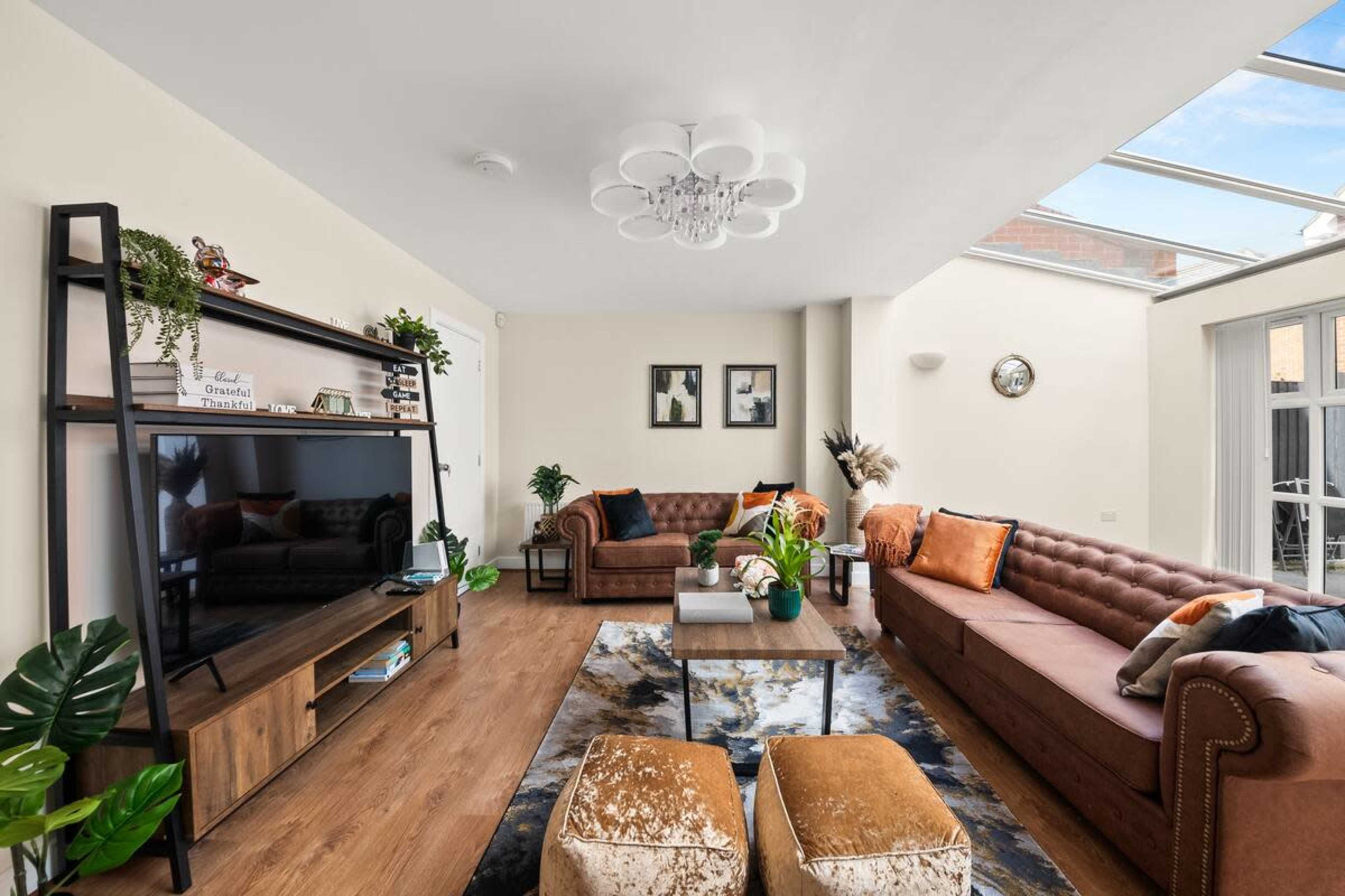 The image shows a bright living room with two brown sofas, a large television, a coffee table in the center, and decorative plants.