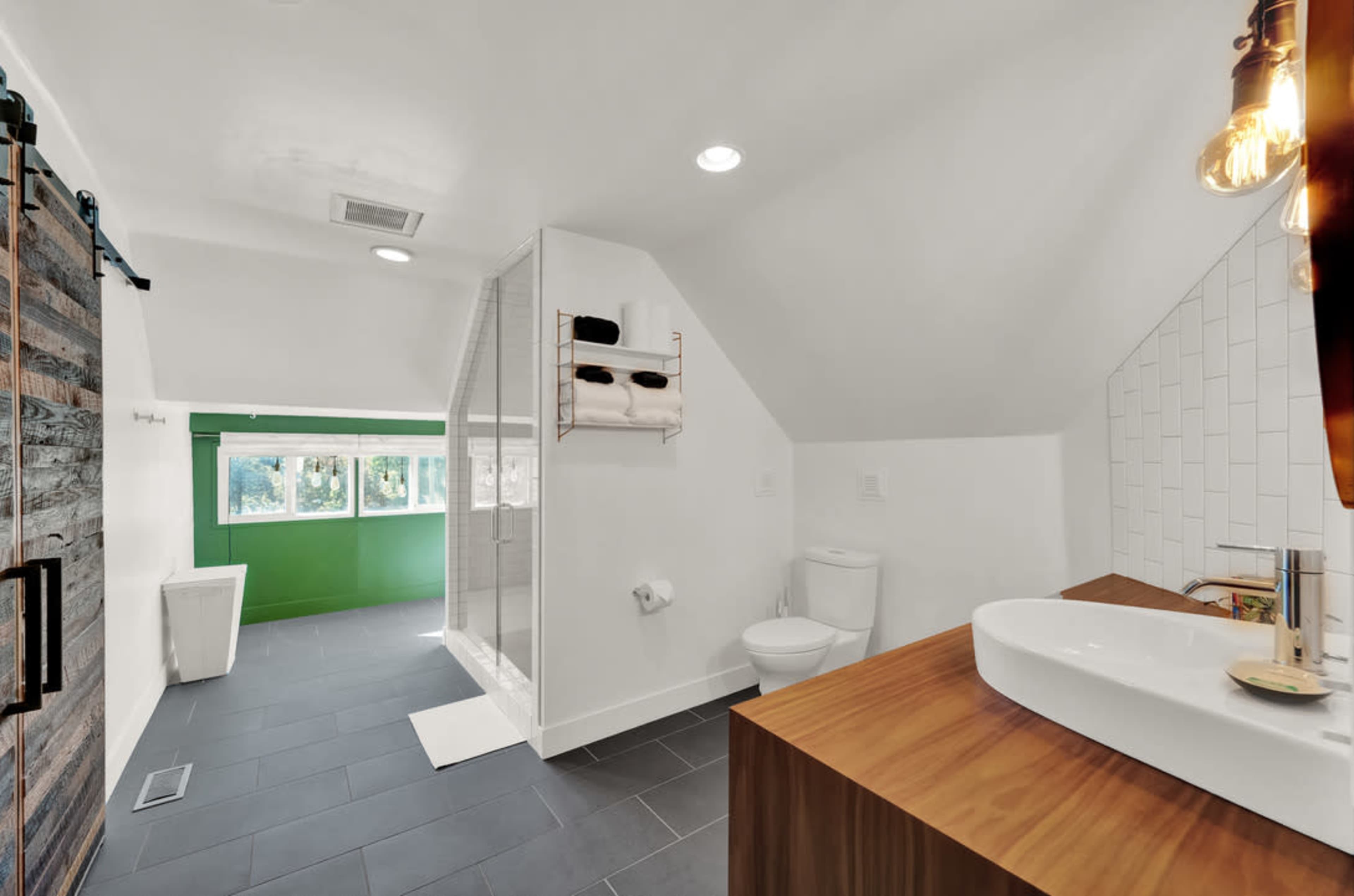 A modern bathroom with a shower, toilet, and a wooden vanity under a sloped ceiling, featuring large windows and a green accent wall.