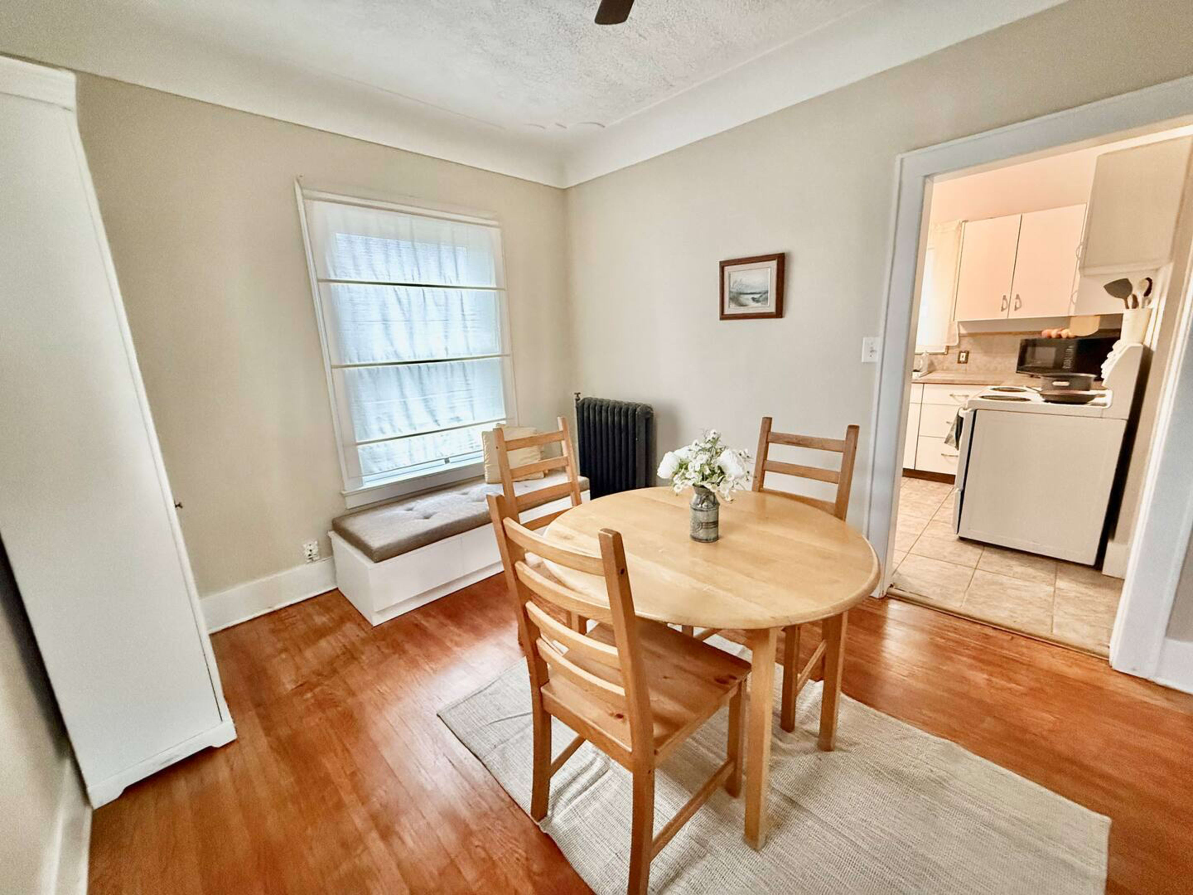 A small dining area features a round wooden table surrounded by four chairs, with a window and a doorway leading to a kitchen.