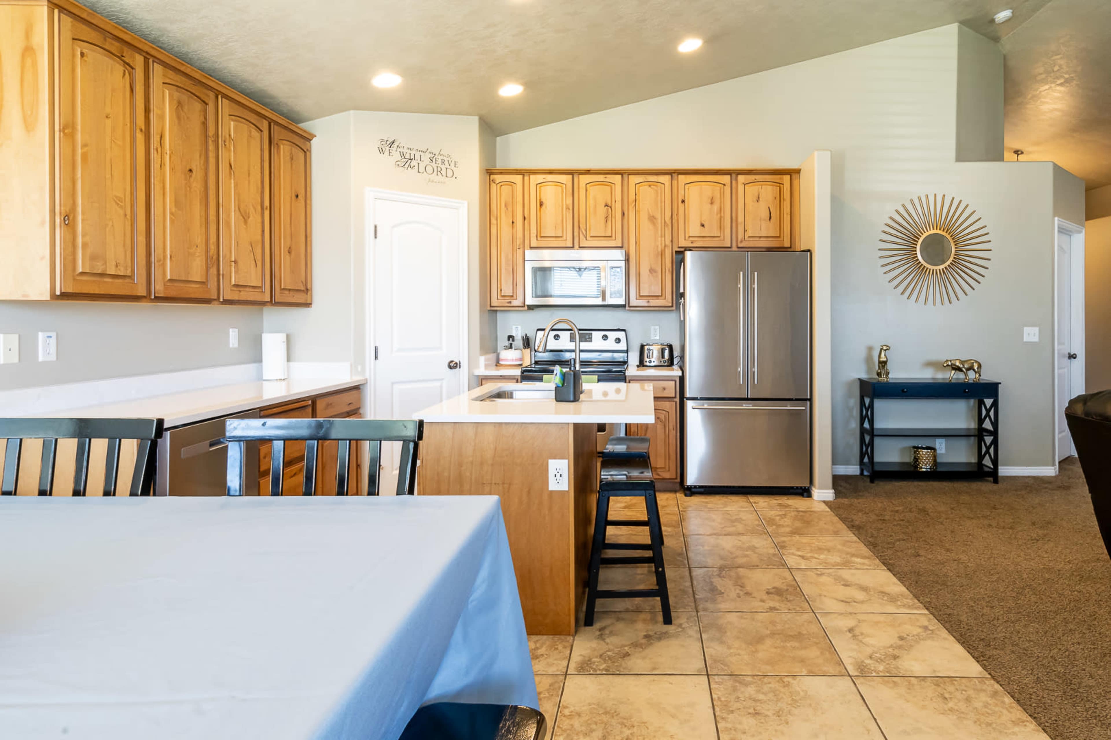 The image shows a modern kitchen with wooden cabinets, a stainless steel refrigerator, and a central island with seating.