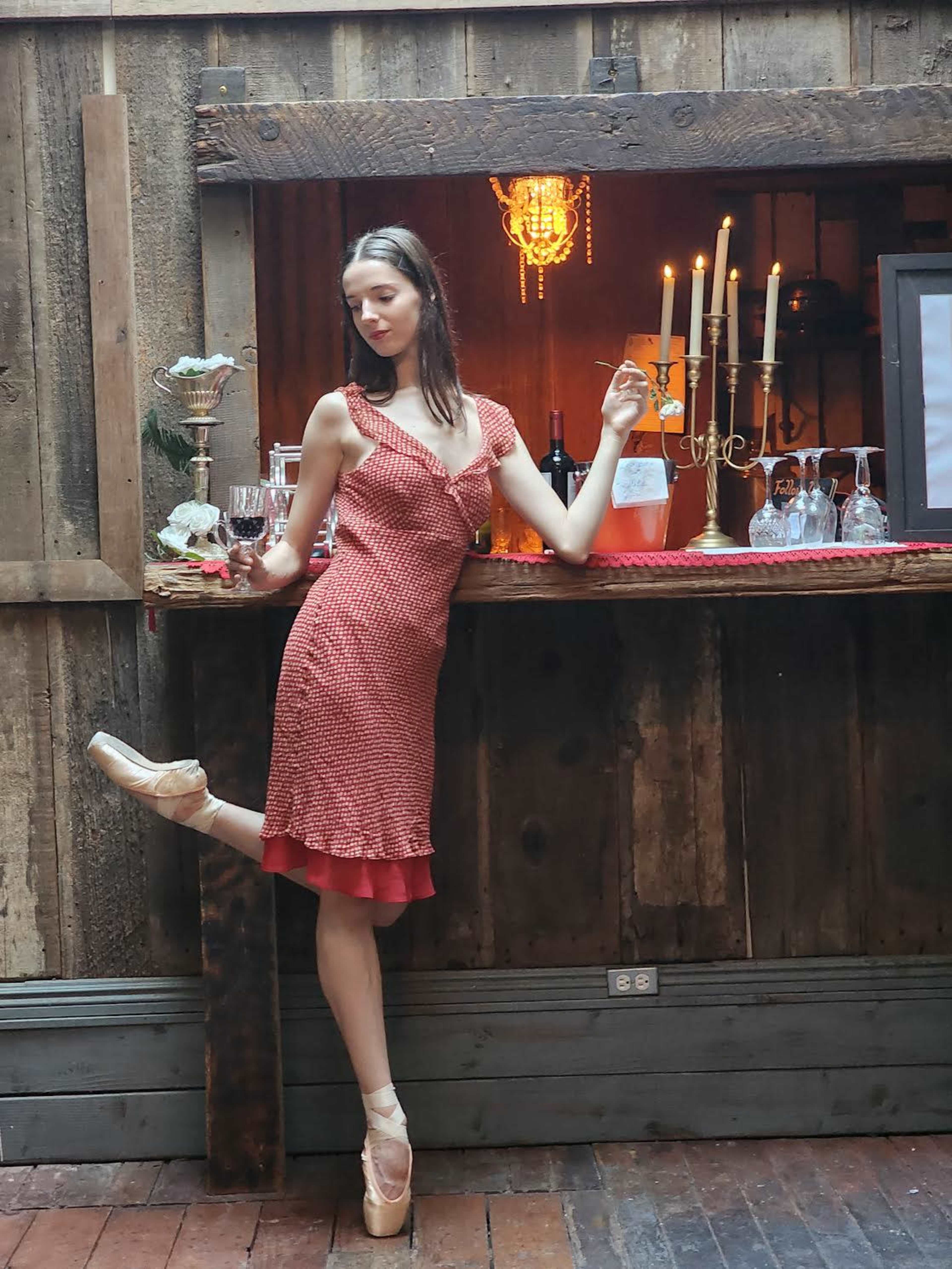 A woman in a red dress poses elegantly by a wooden bar adorned with candles and glassware.
