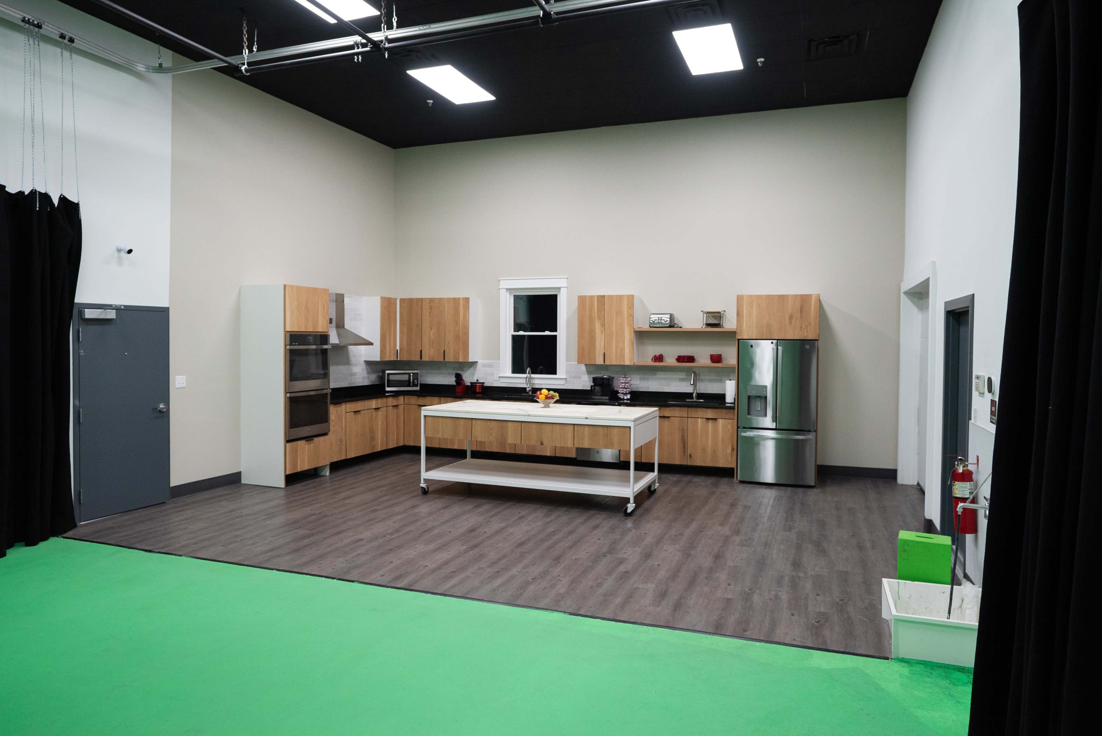 The image shows a spacious kitchen setup with modern appliances, wooden cabinetry, and a central island under bright lighting in a studio environment.