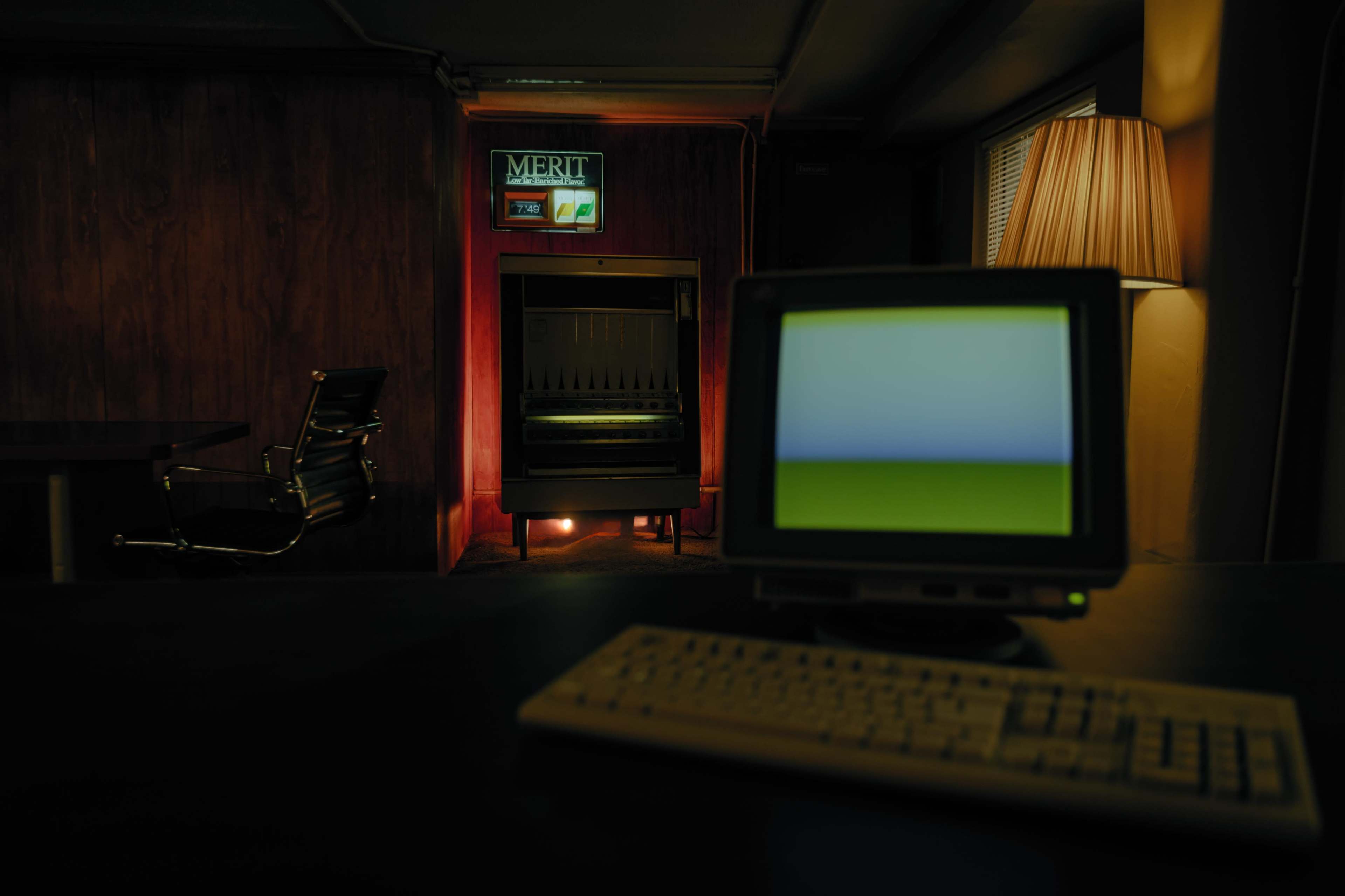 A dimly lit room with a retro computer and keyboard in the foreground, while a neon sign displaying "MERIT" and a darkened fireplace are visible in the background.