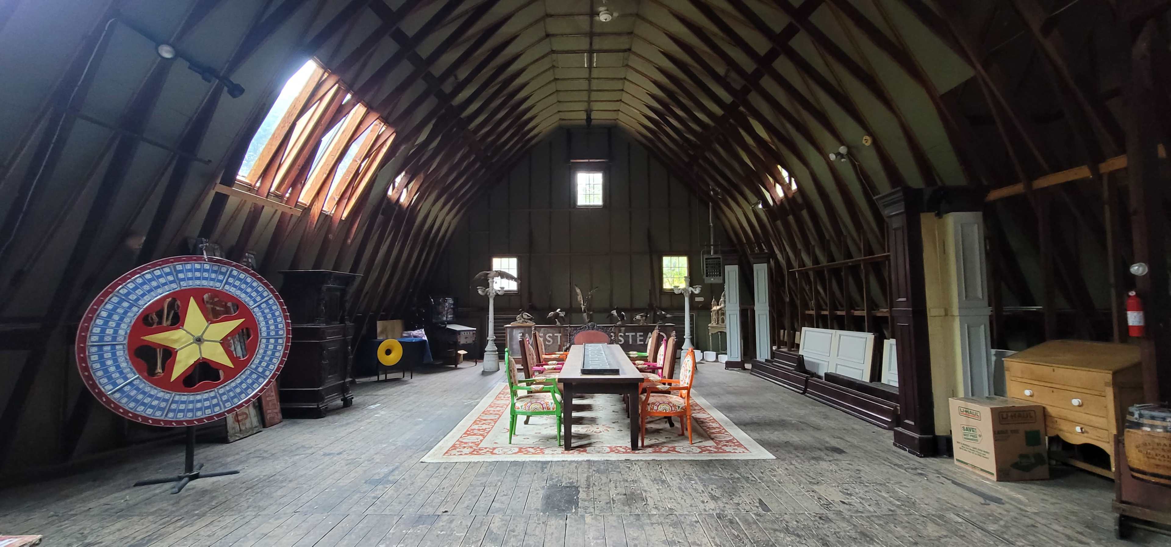 A spacious attic with wooden beams, featuring a long dining table surrounded by brightly colored chairs, an ornate circular decoration on the left, and various pieces of furniture scattered around.