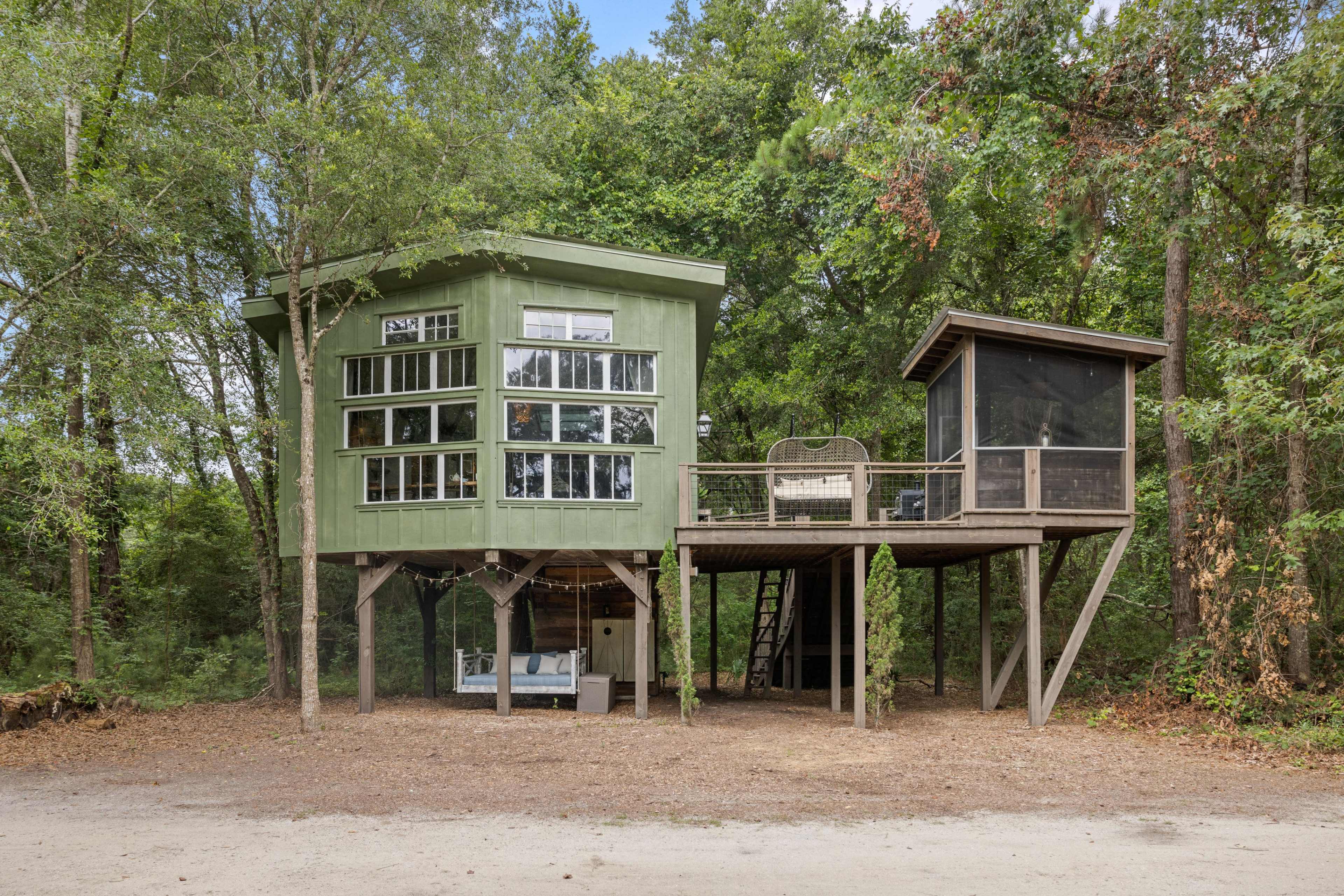 A modern green treehouse with large windows, elevated on stilts, adjacent to a smaller screened porch.
