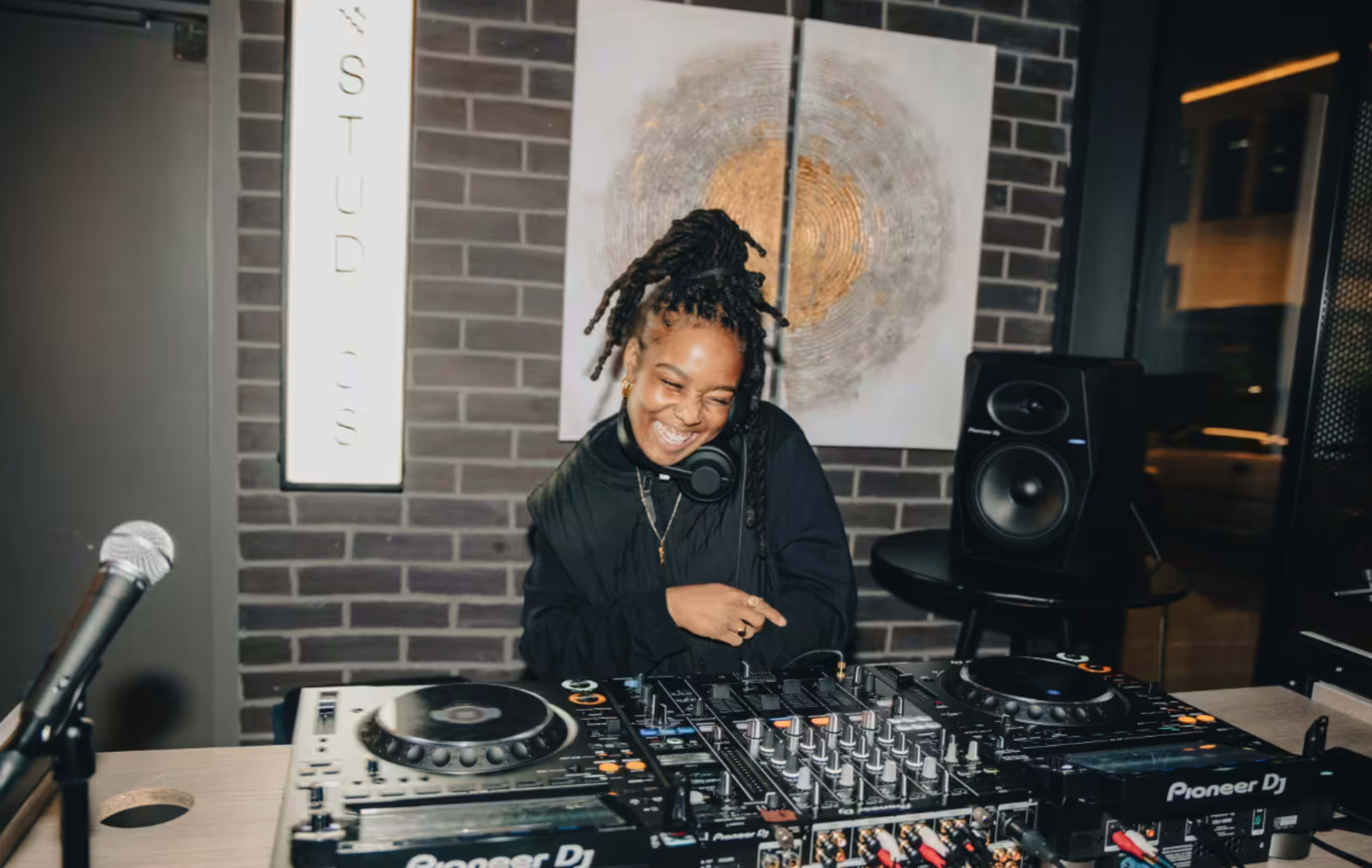 A DJ smiles while mixing music at a professional setup in a studio featuring a brick wall and speakers.