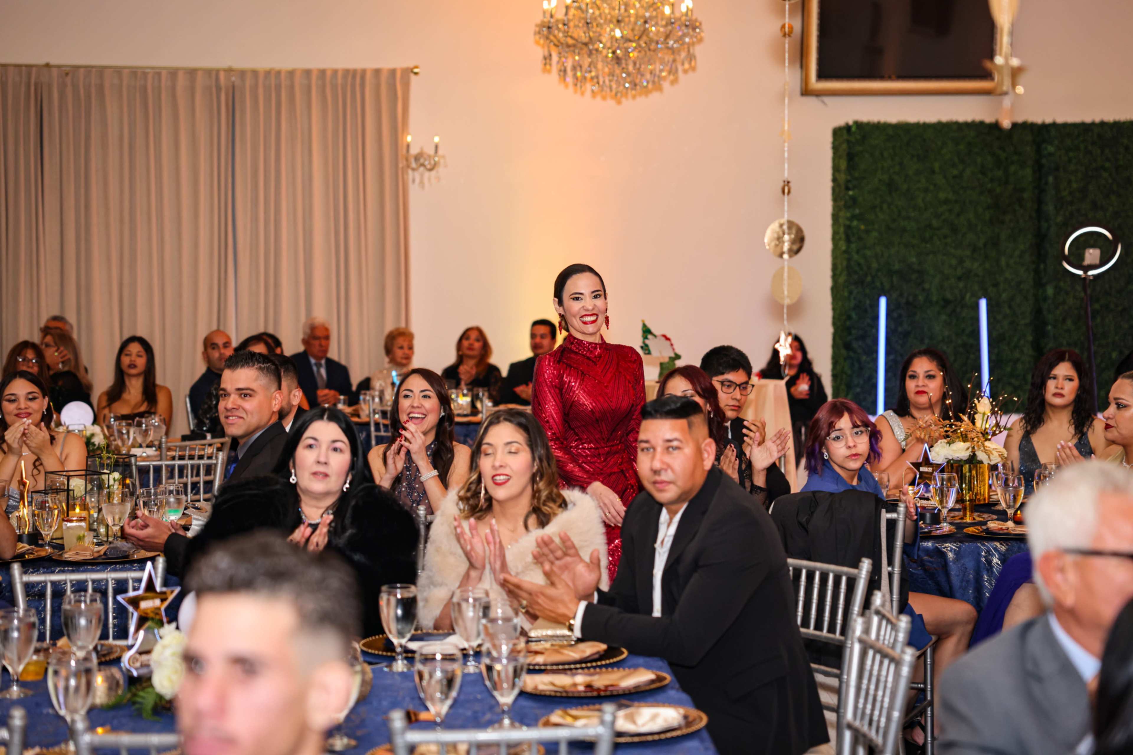 A group of elegantly dressed guests claps while seated at round tables adorned with decorations during a celebratory event in a banquet hall.