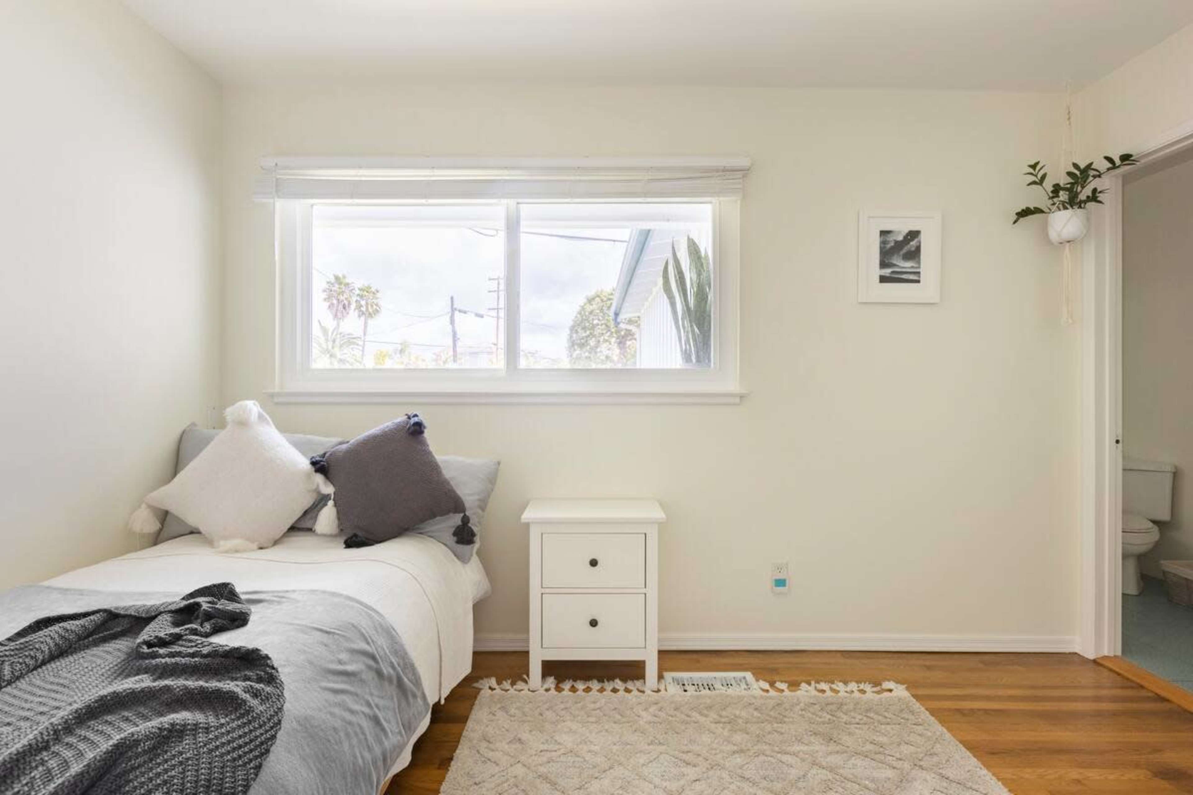 A simple bedroom with a single bed covered in gray and white bedding, a small nightstand beside it, and a window allowing natural light to enter.