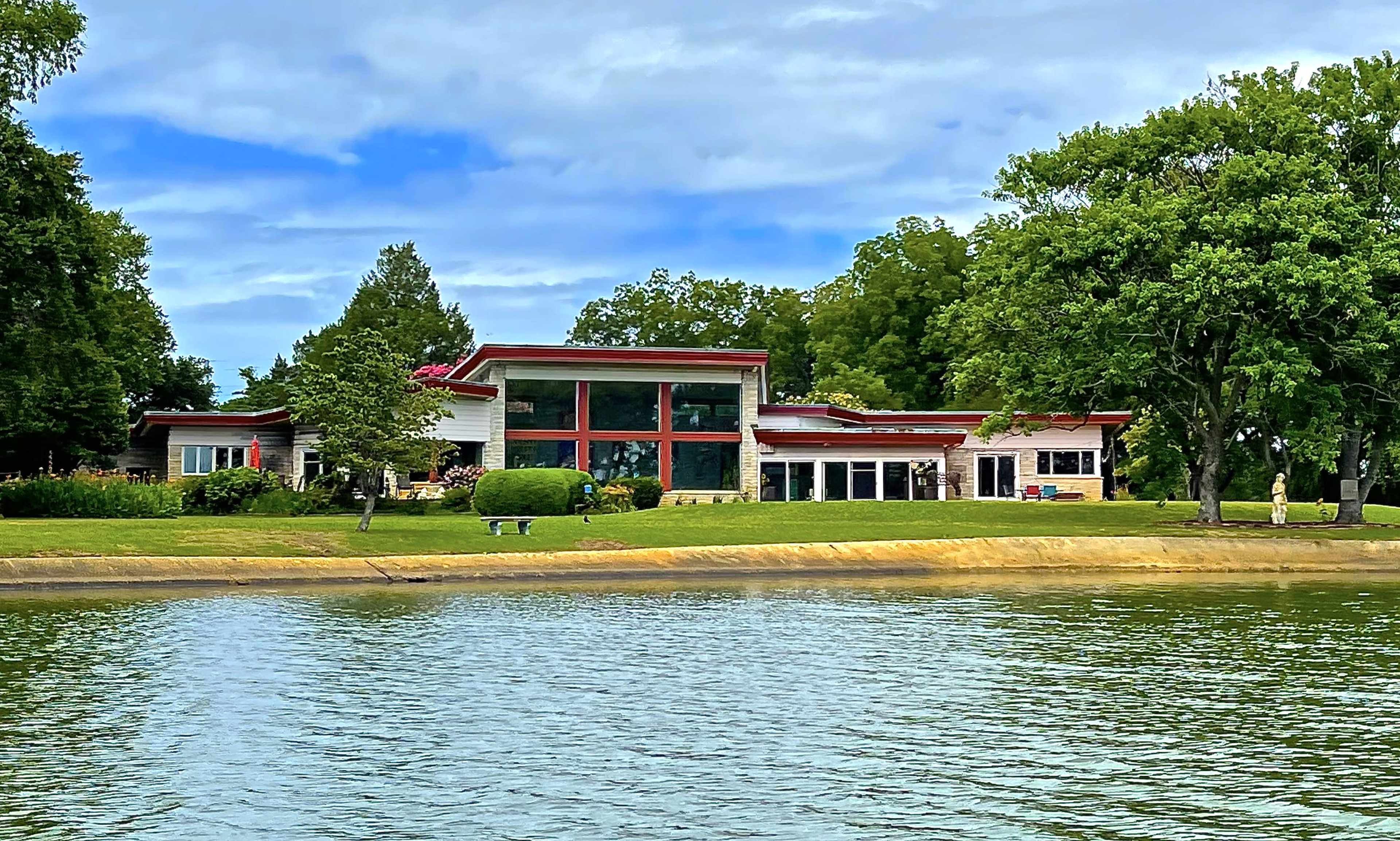 A large modern house with a red roof and glass windows is situated along the shoreline of a lake, surrounded by trees and greenery.