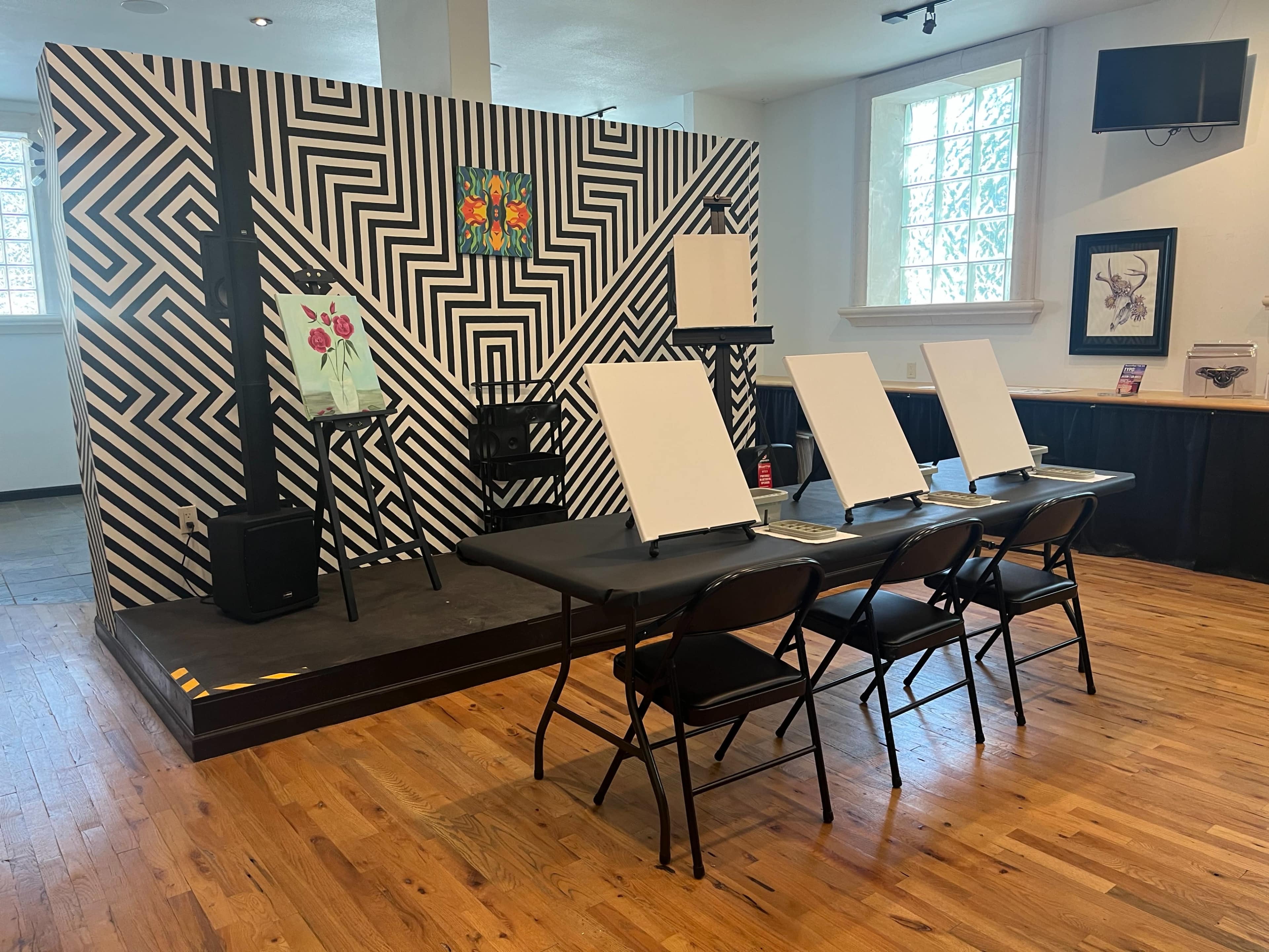 The image shows a room with a stage featuring a black and white striped backdrop, four empty easels in front of a table with chairs, and a painting on display.