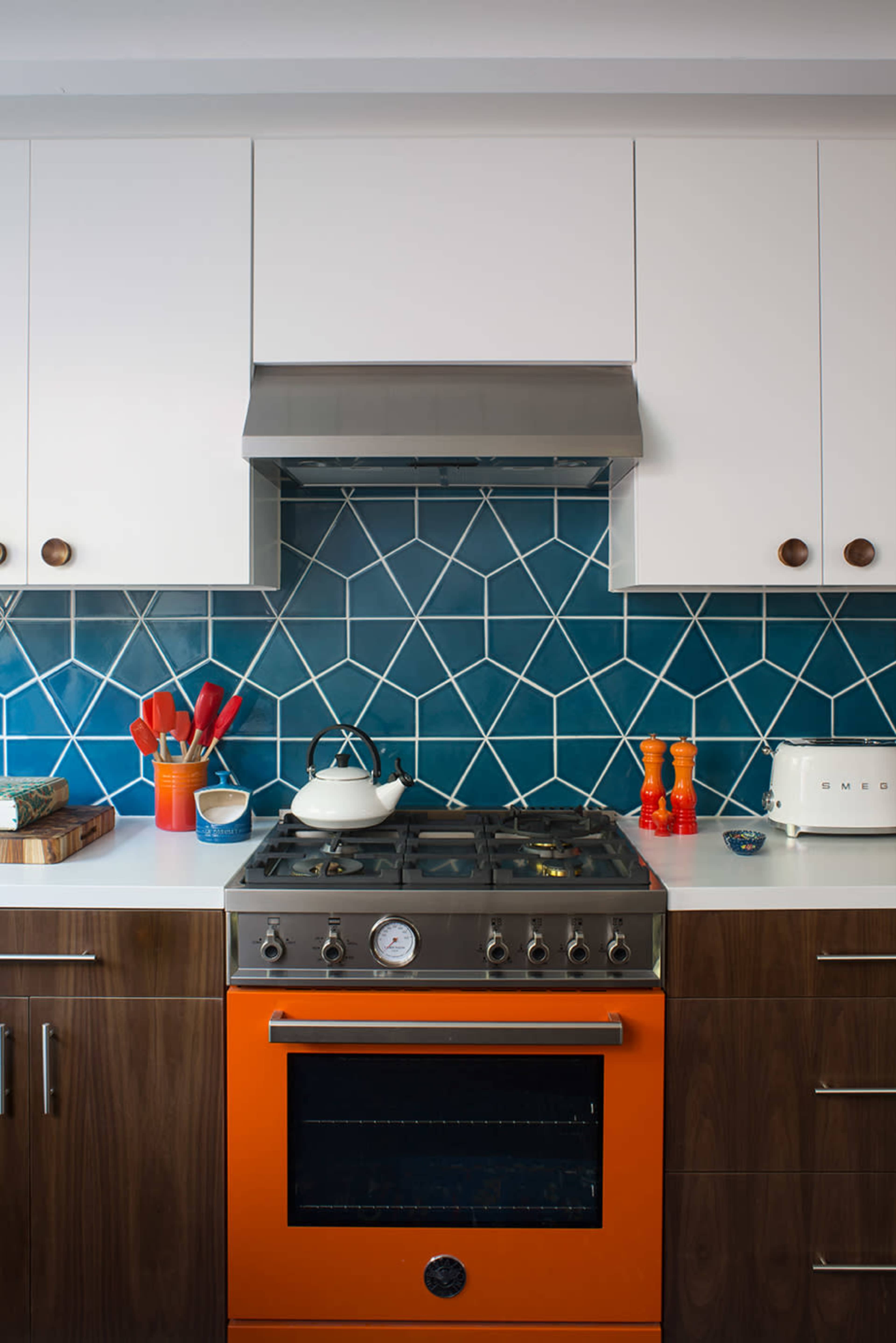 The kitchen features an orange stove beneath a stainless steel hood and blue geometric backsplash, complemented by white cabinets and wooden drawers.