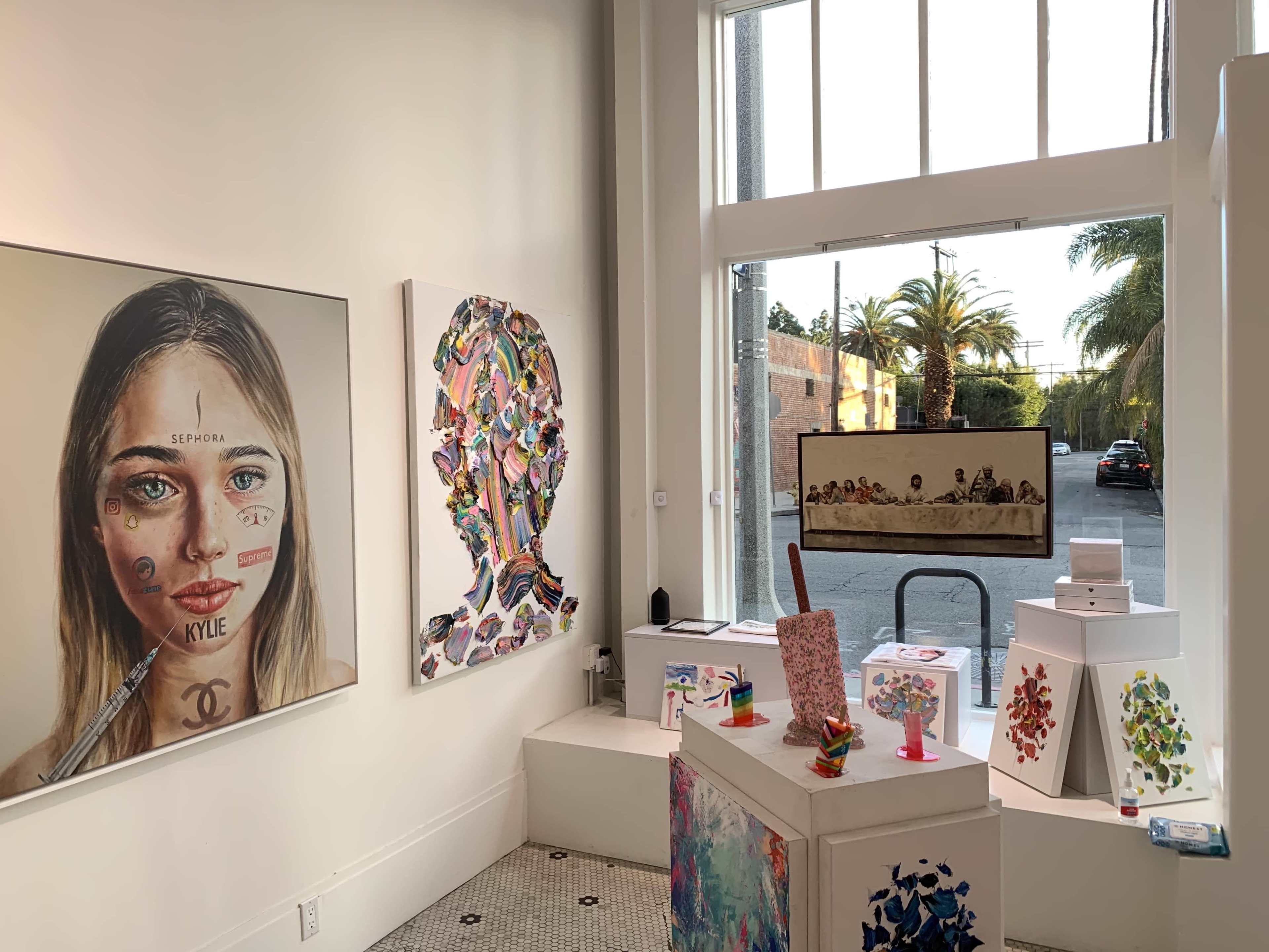 An art gallery with two prominent artworks on the walls, one of a portrait and the other abstract, alongside a display area featuring colorful sculptures and a view of palm trees outside the window.