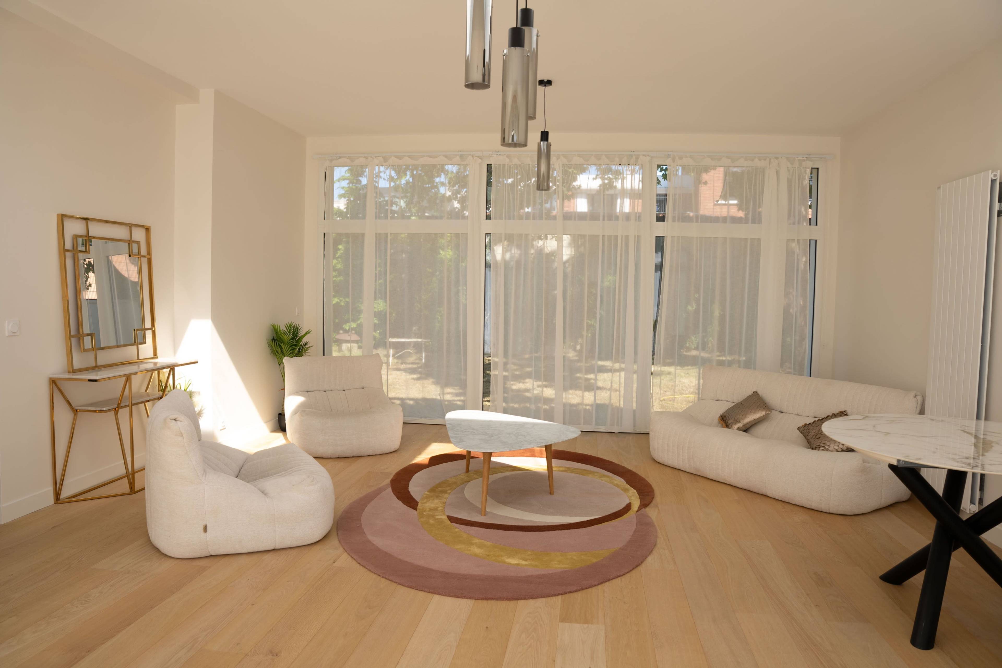 A modern living room features a minimalistic design with a circular area rug, cozy seating, a coffee table, and large windows allowing natural light to enter.