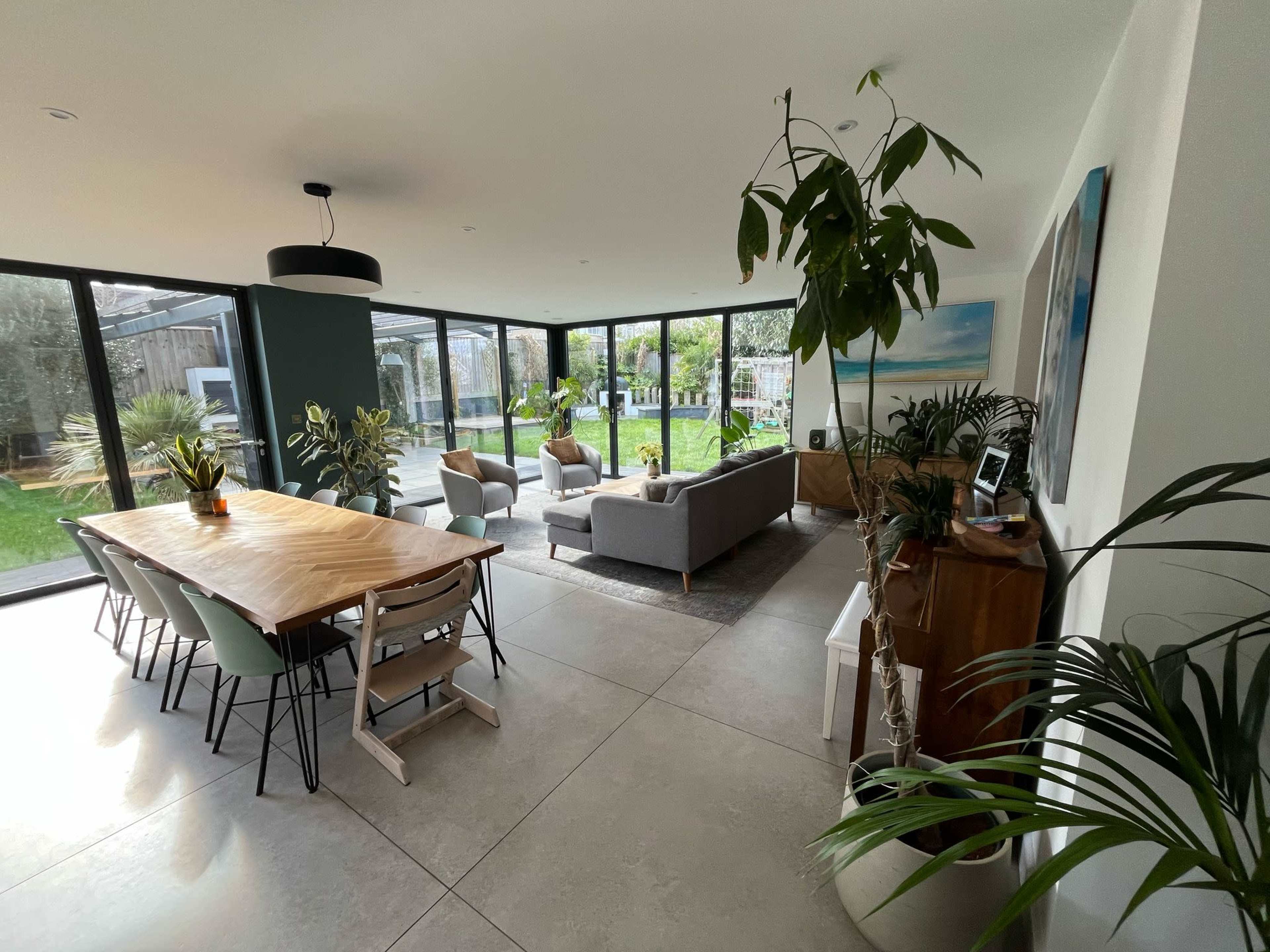 The image shows a spacious, modern living area with large windows, a wooden dining table, a gray sofa, and various indoor plants.