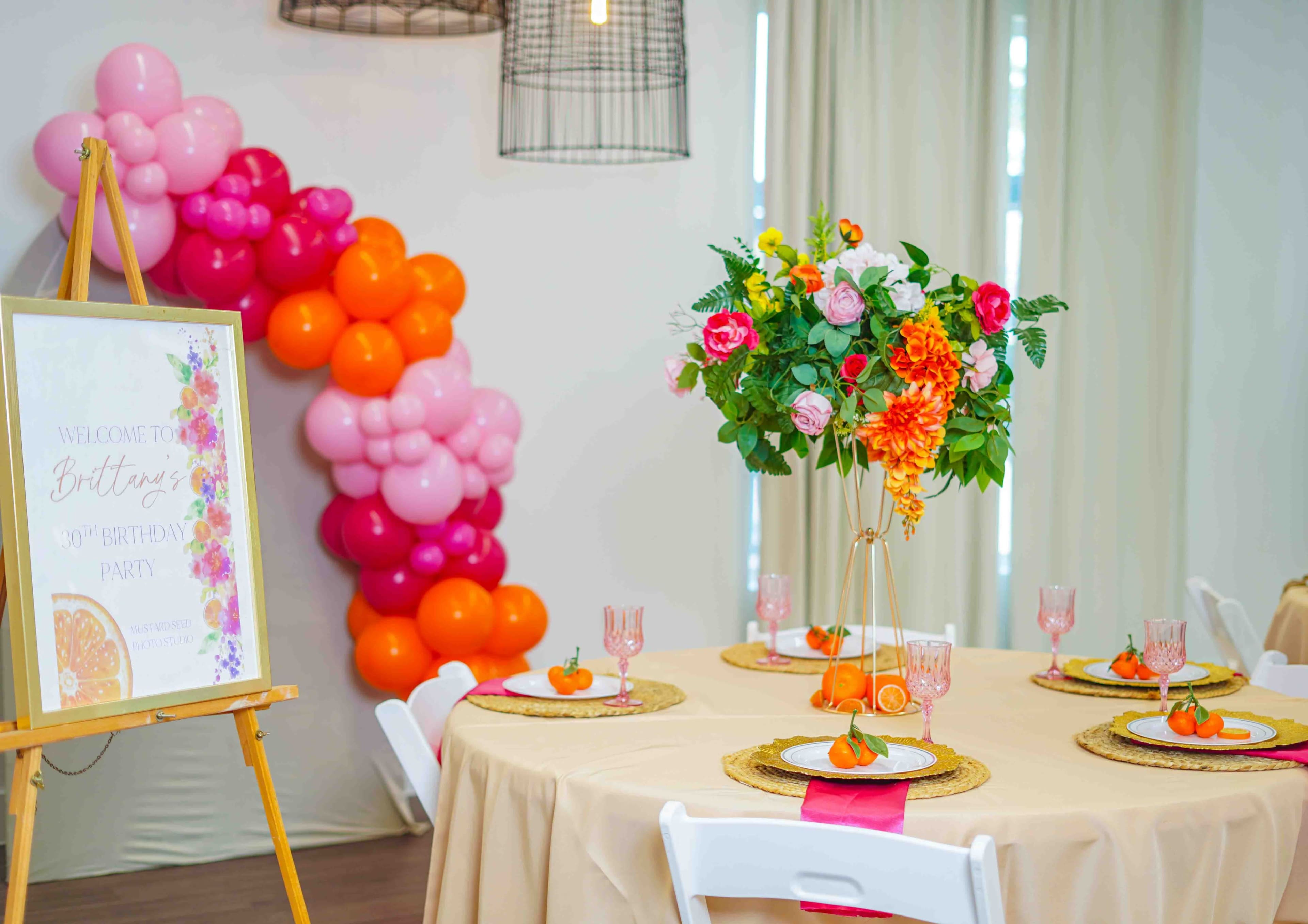 The image shows a decorated table set for a birthday party, featuring a floral centerpiece, colorful balloons in the background, and a welcome sign.