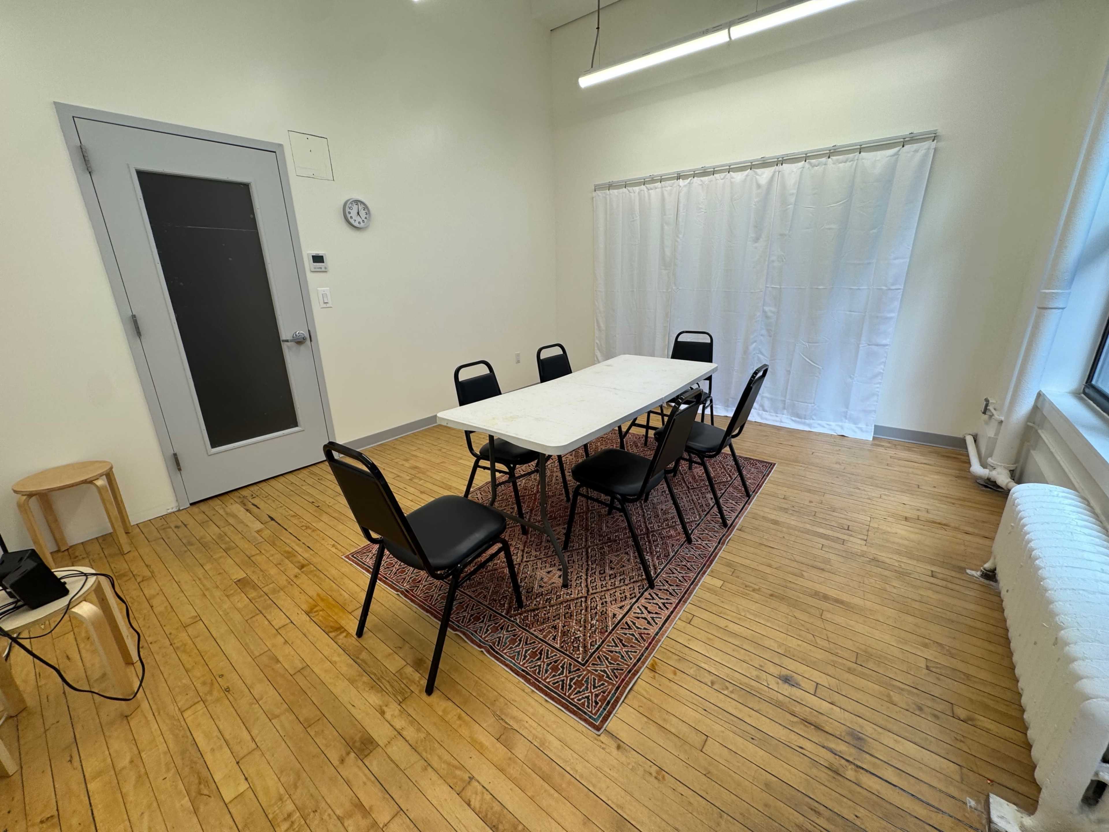 The image shows a simple meeting room with a rectangular table surrounded by six chairs, a stool in the corner, a wall clock, and a curtain covering a window.