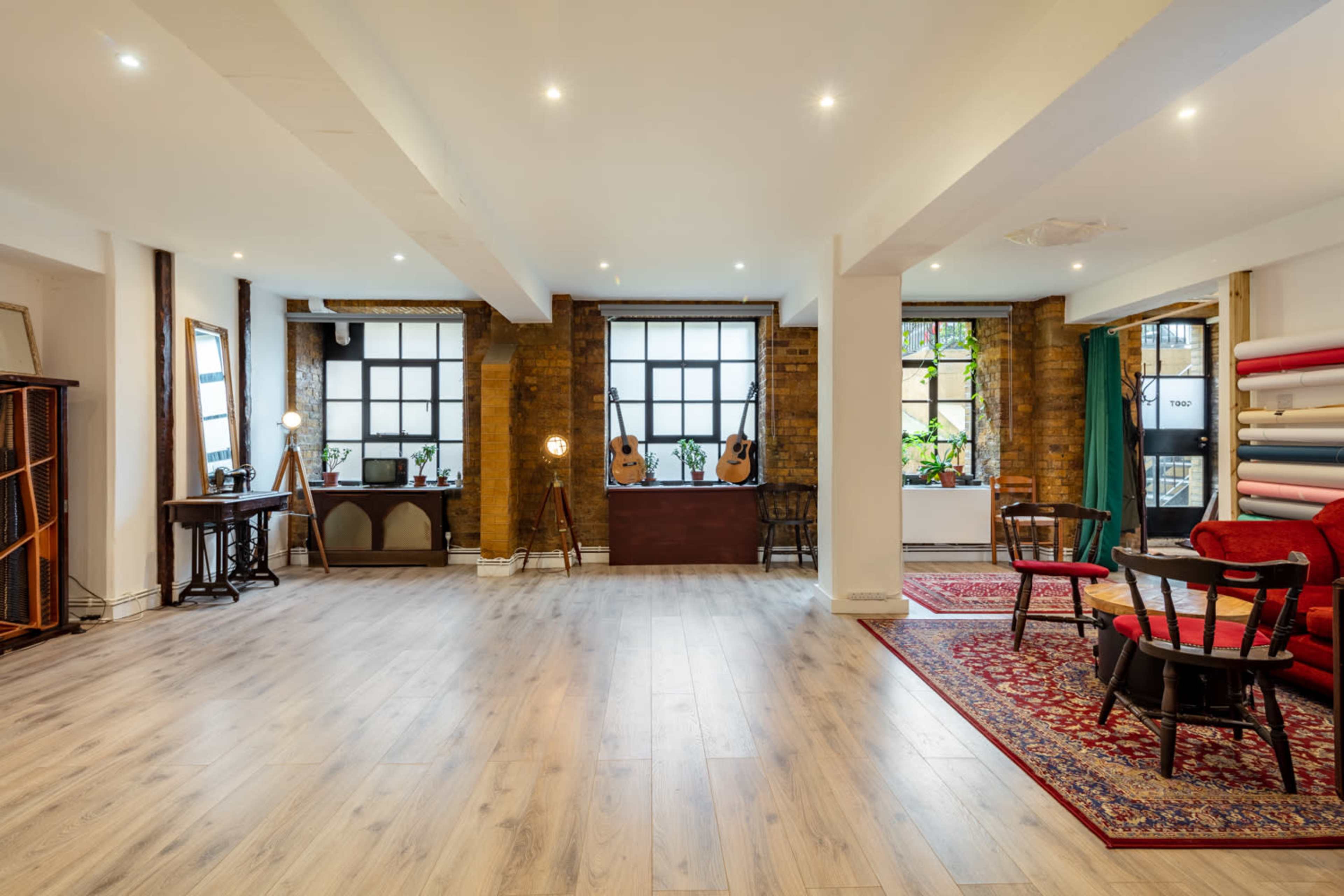 A spacious room with wooden flooring, large windows, and various furniture pieces including chairs, a rug, and decorative items.