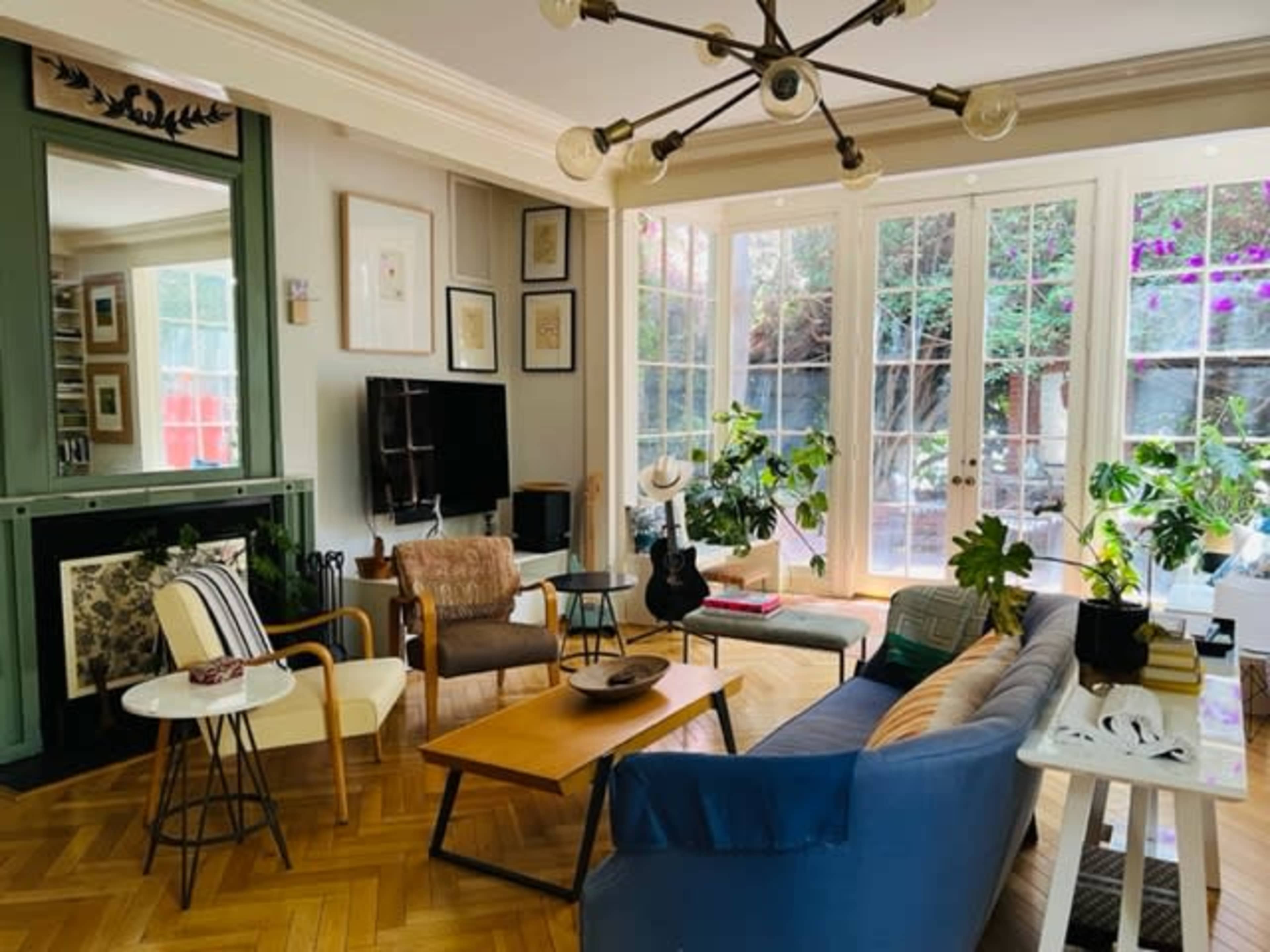 The living room features a mix of modern and vintage furniture, large glass doors leading to a garden, and several indoor plants.