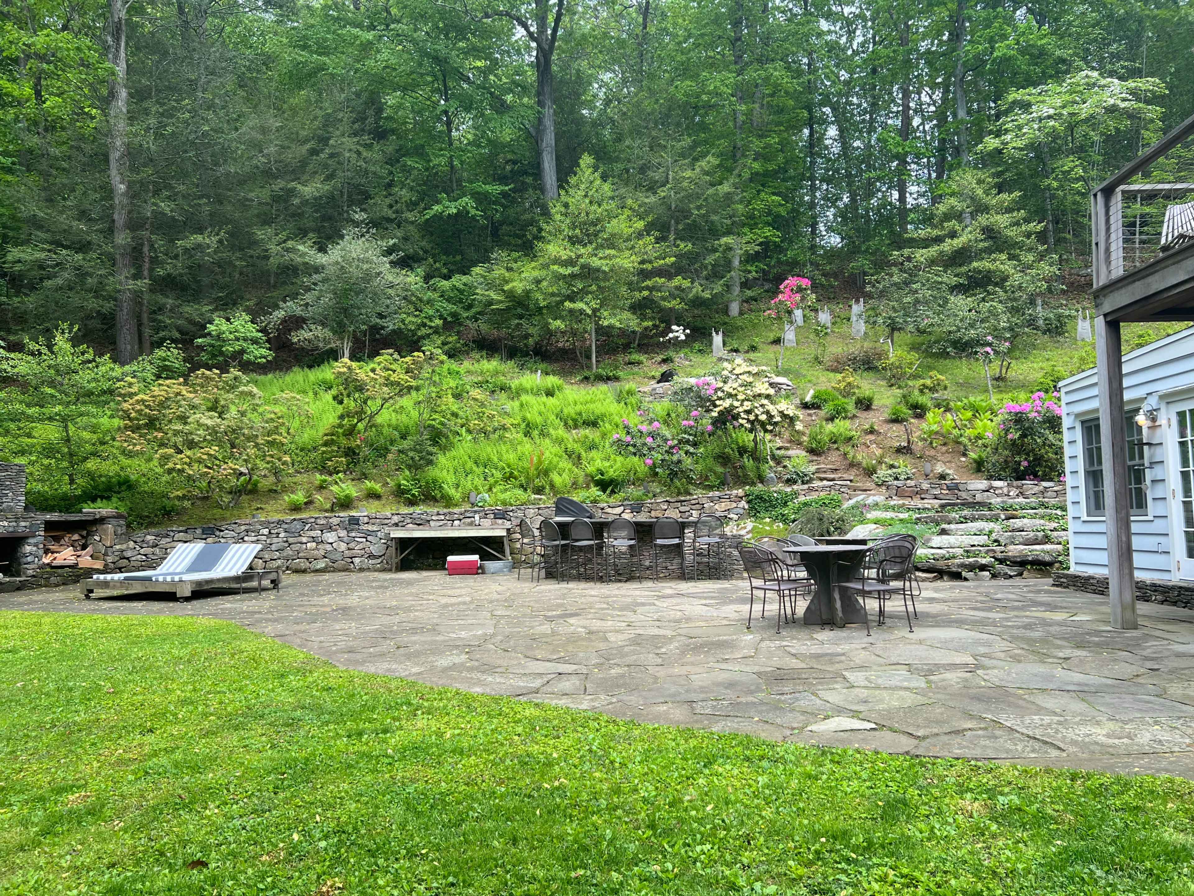 A stone patio with a table and chairs is surrounded by lush greenery and landscaping features, including a fire pit and two lounge chairs.