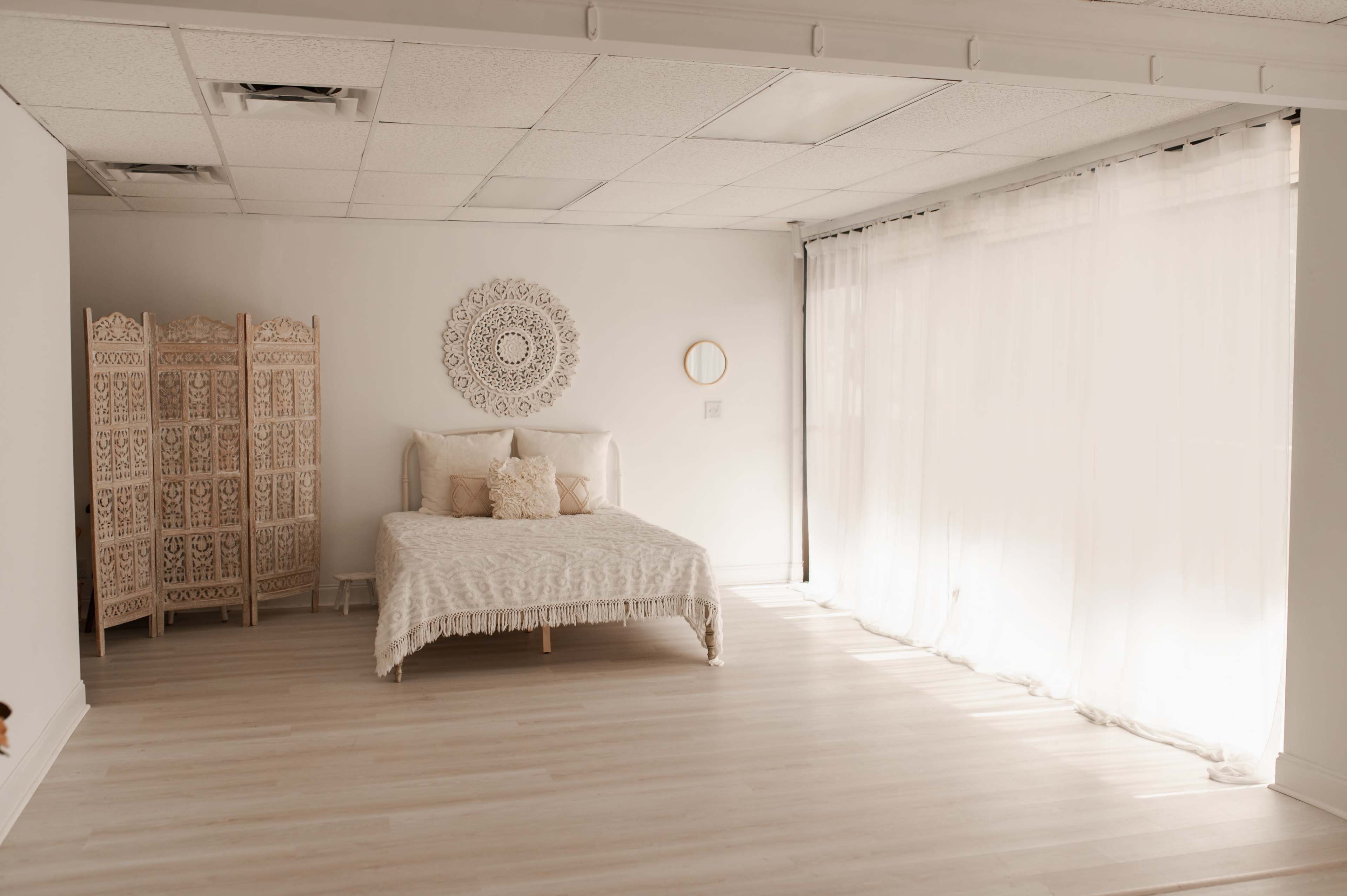 The image depicts a minimalist bedroom featuring a bed with light bedding, a decorative wall piece, and large windows covered with sheer curtains allowing natural light to fill the space.