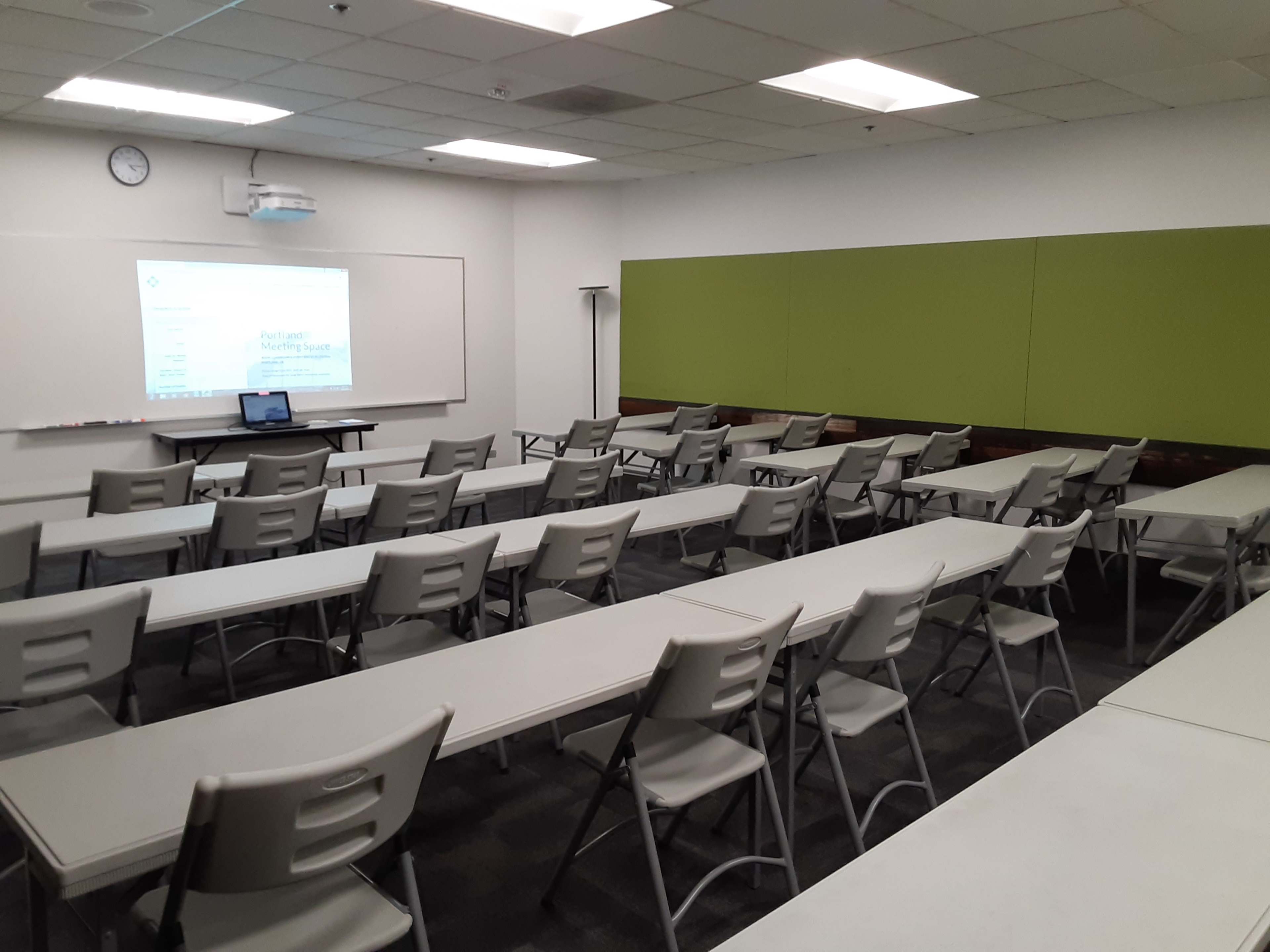 A classroom arranged with several rows of tables and chairs, facing a wall-mounted screen and projector.
