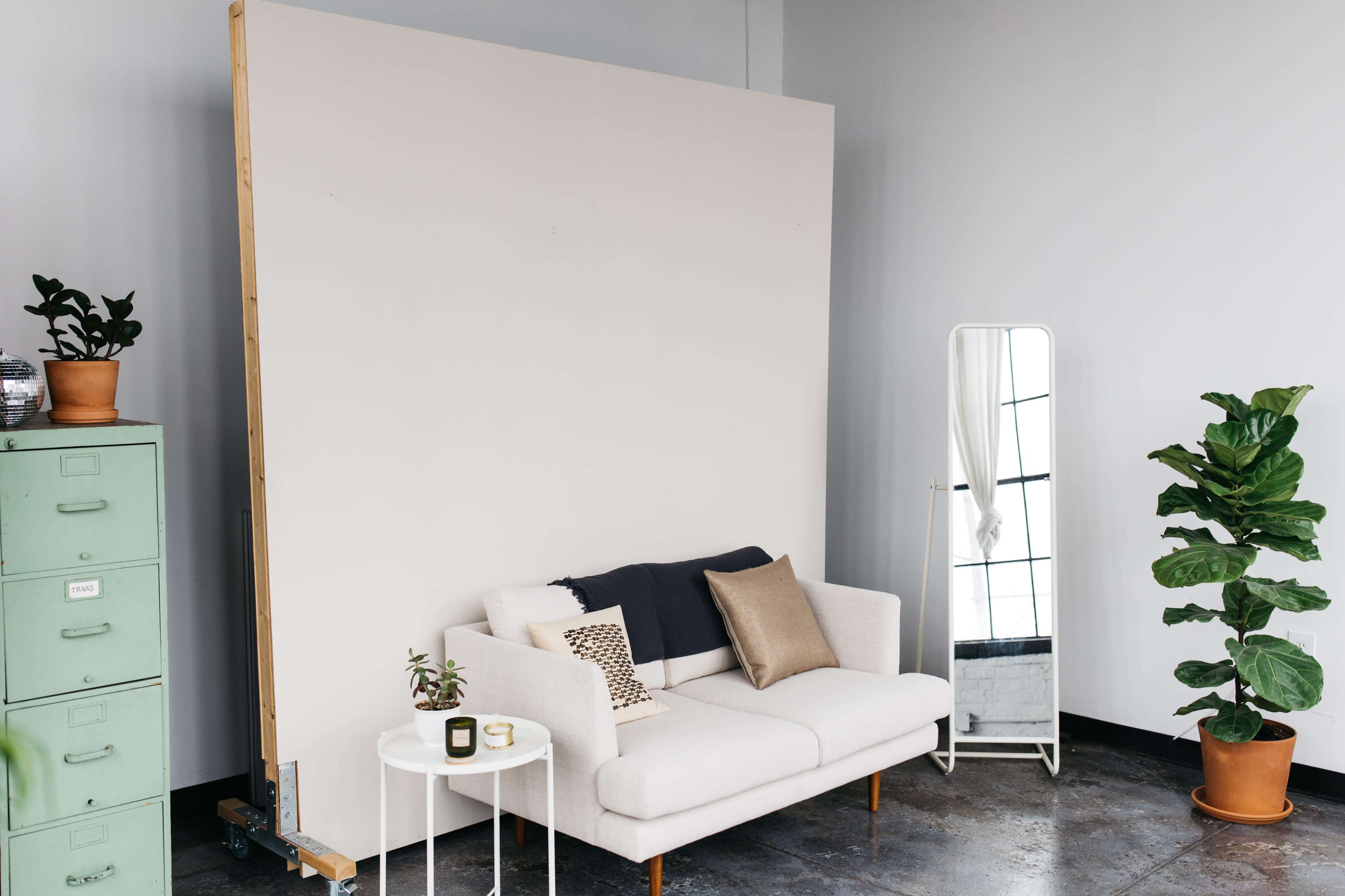 The image shows a minimalist interior with a white sofa, a side table with a candle, a large blank wall, a full-length mirror, and potted plants.
