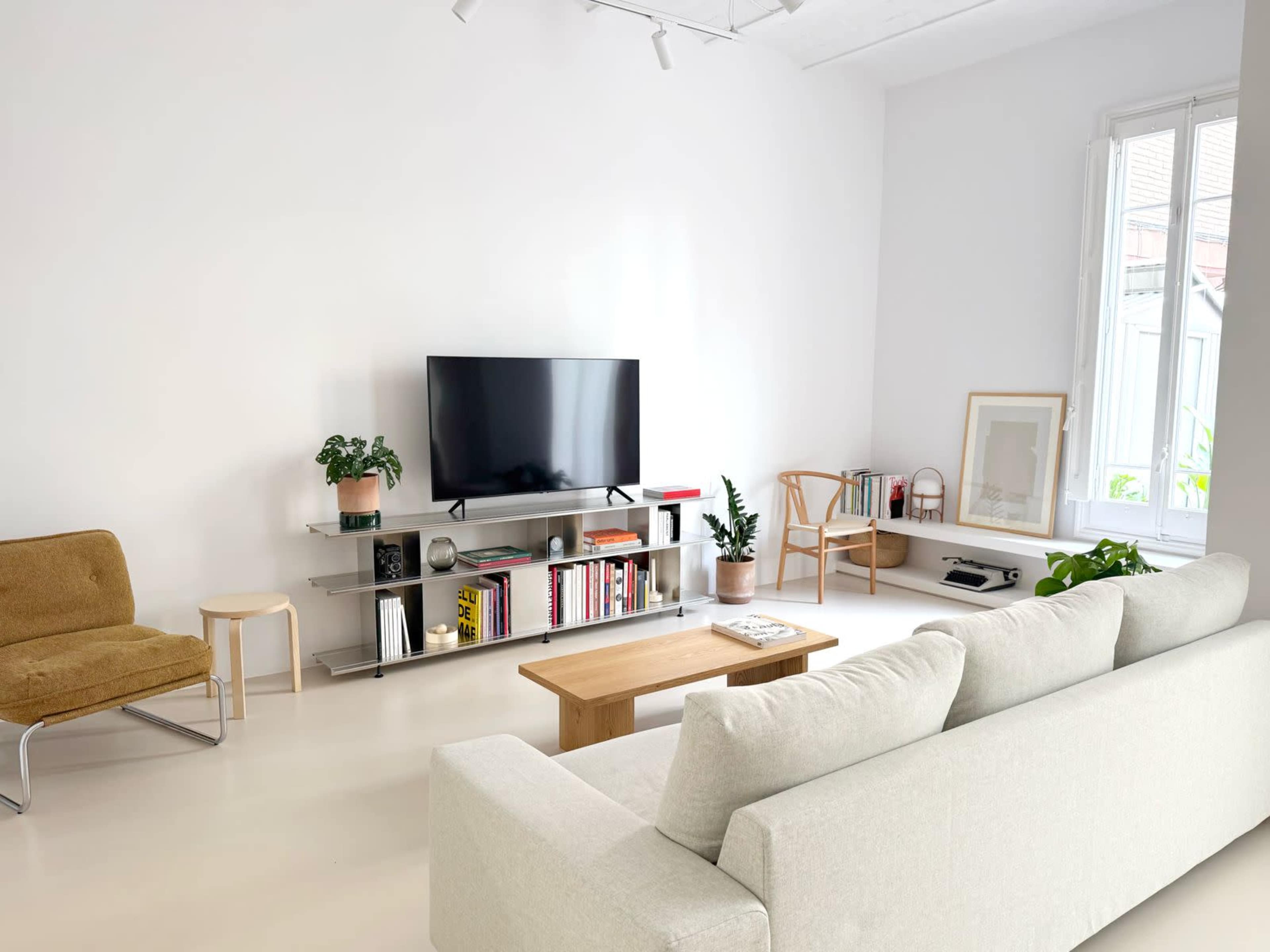 The image shows a modern, minimalistic living room with a light-colored sofa, a television on a sleek shelf, and plants in the corner.
