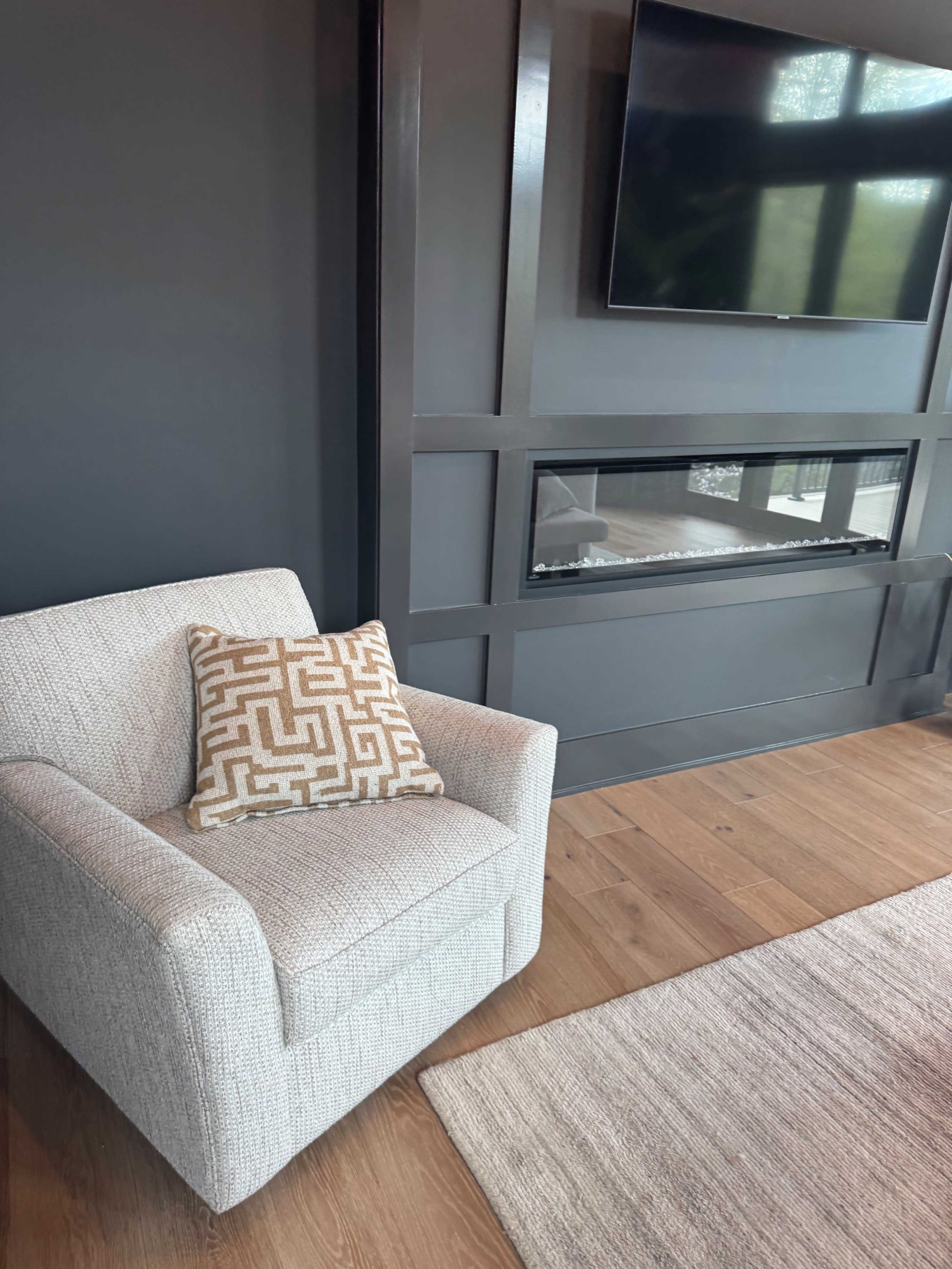 The image shows a cozy living room corner with a light-colored armchair, a decorative pillow, a mounted television, and a modern electric fireplace beneath a dark wall.