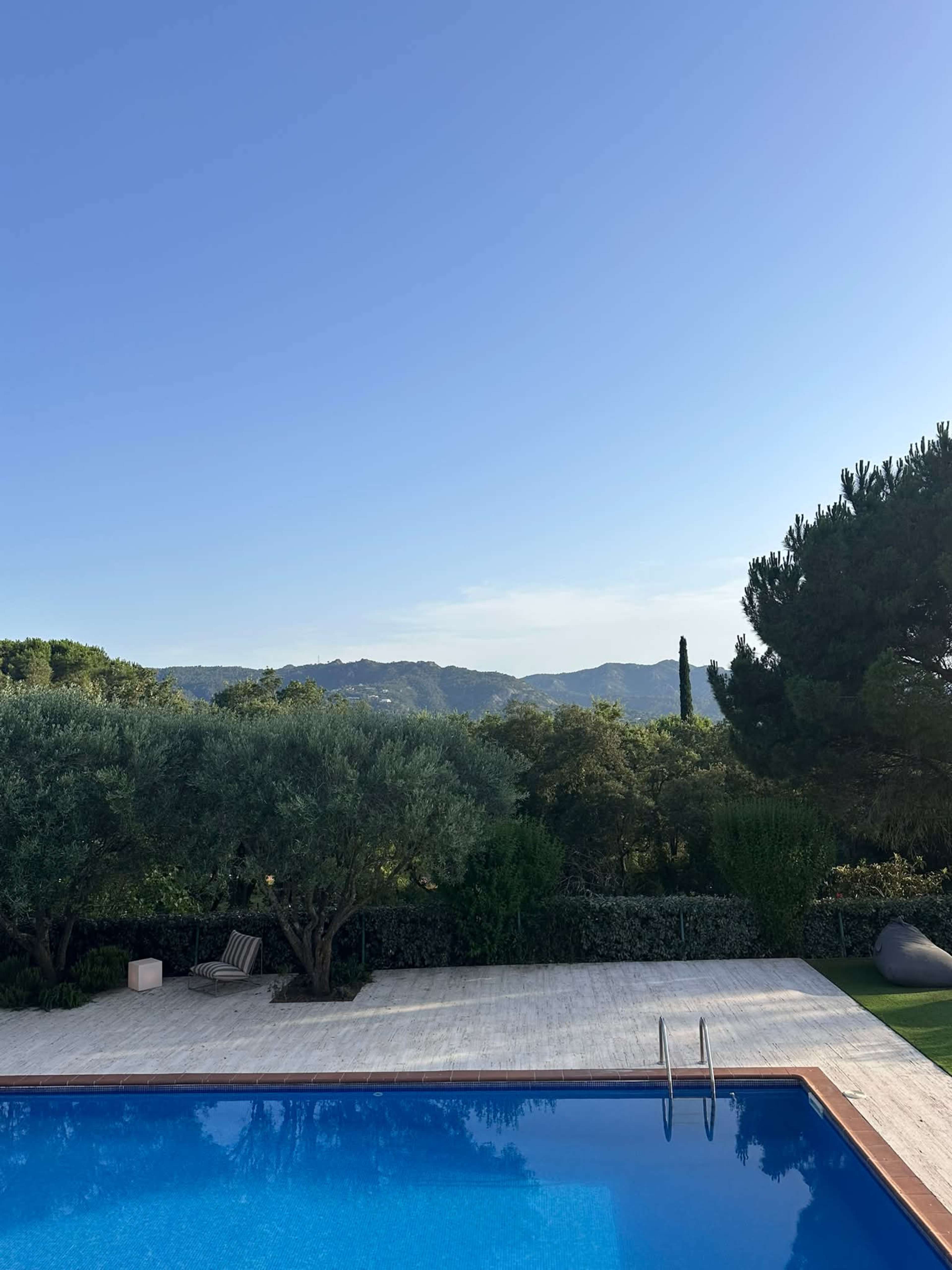 A clear blue sky overlooks a swimming pool surrounded by greenery and hills in the background.
