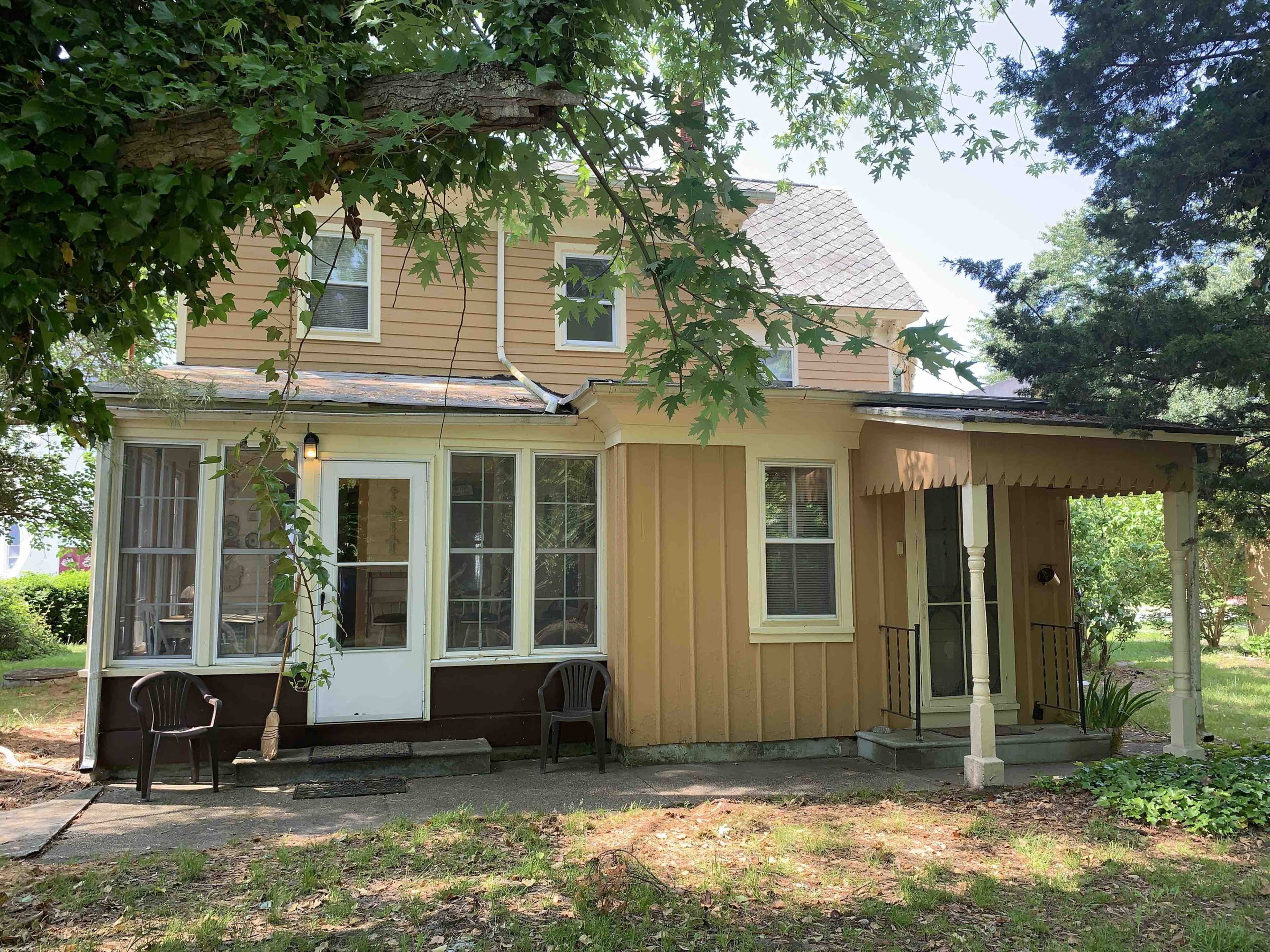 A two-story house with brown and tan siding features a front porch and large windows, surrounded by trees and a grassy area.