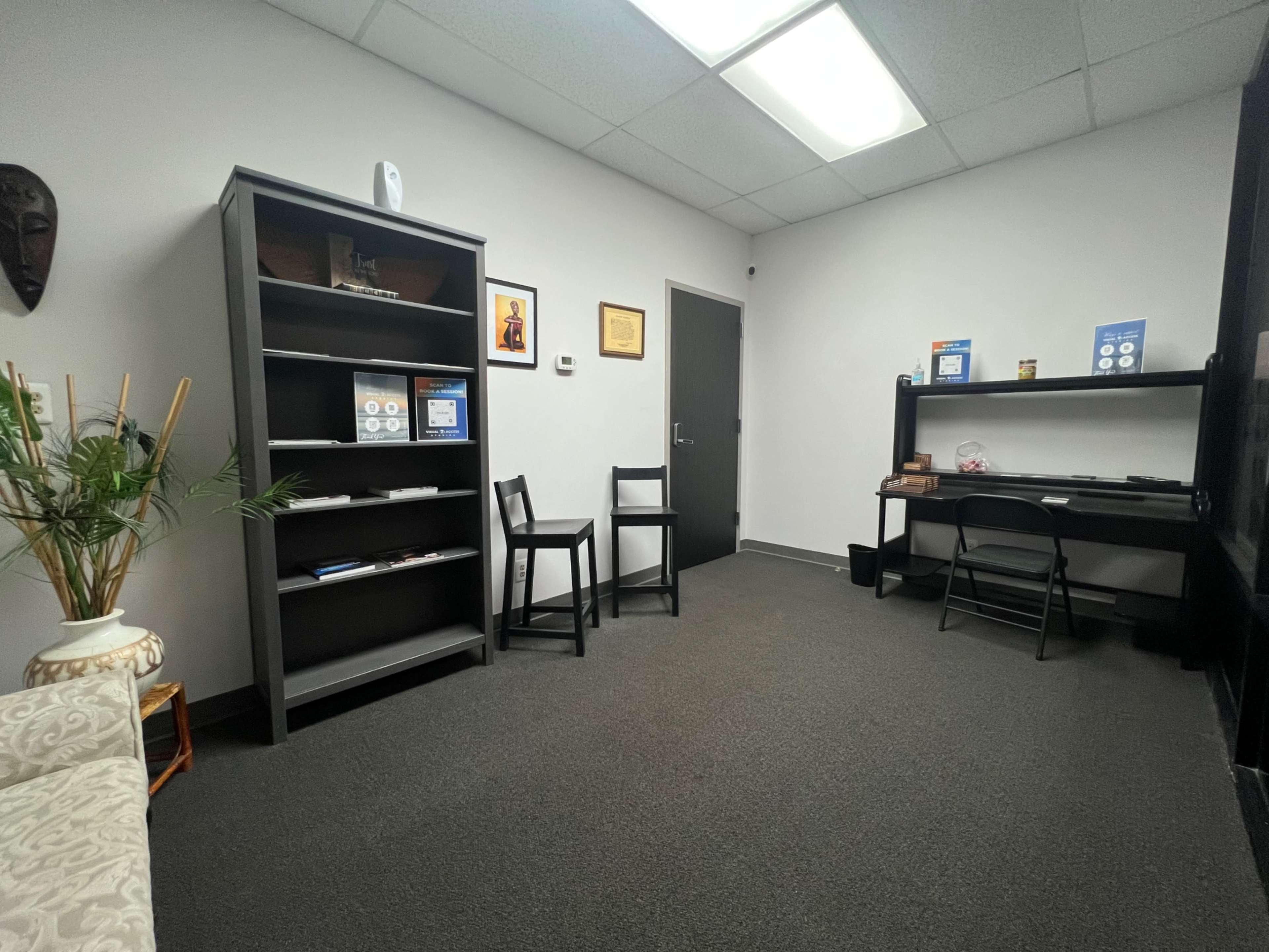 A small, sparse office space with a bookshelf, two chairs, and a desk against the walls.