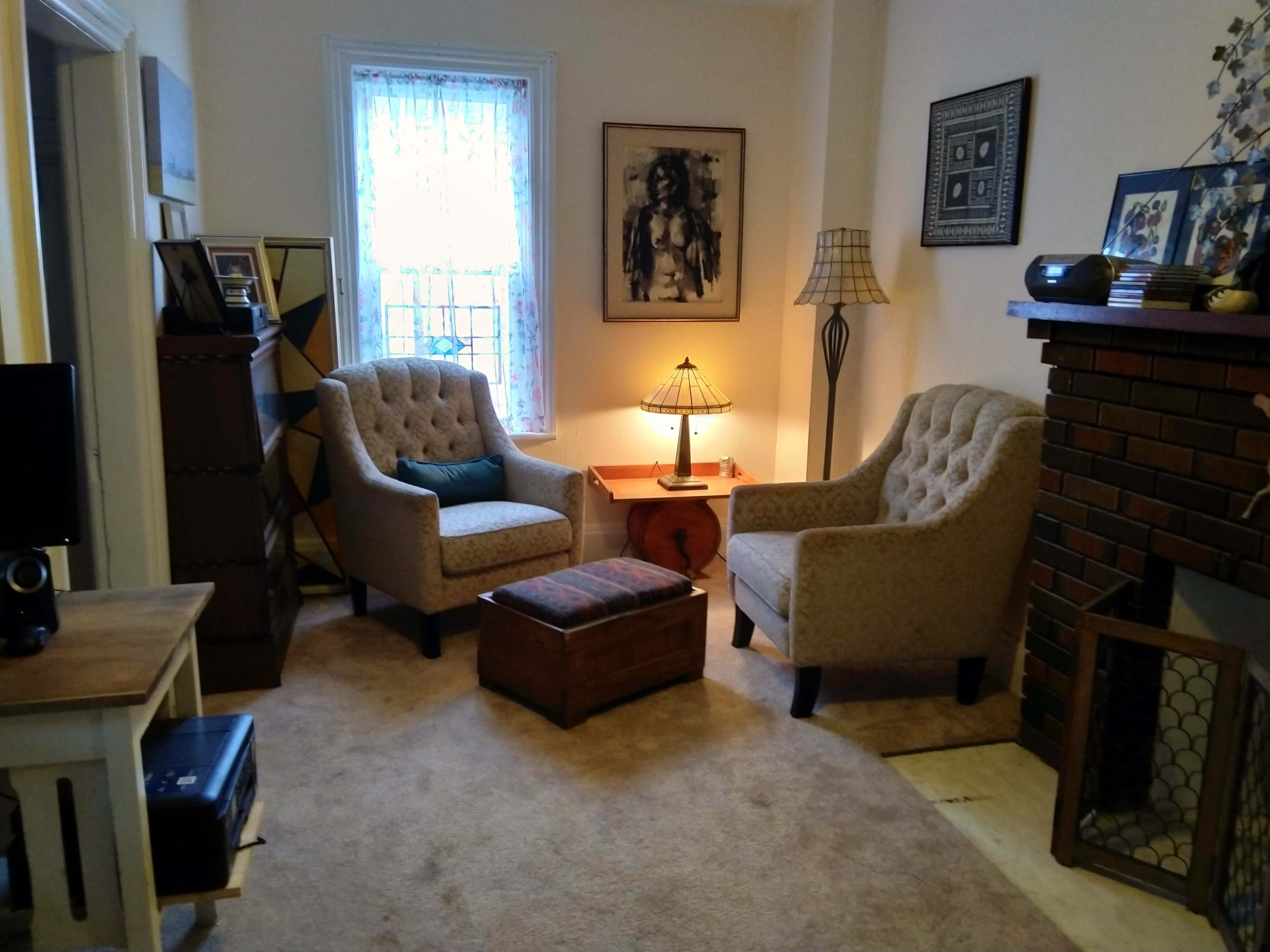 A cozy living room with two upholstered chairs, a small wooden table with a lamp, and a brick fireplace.