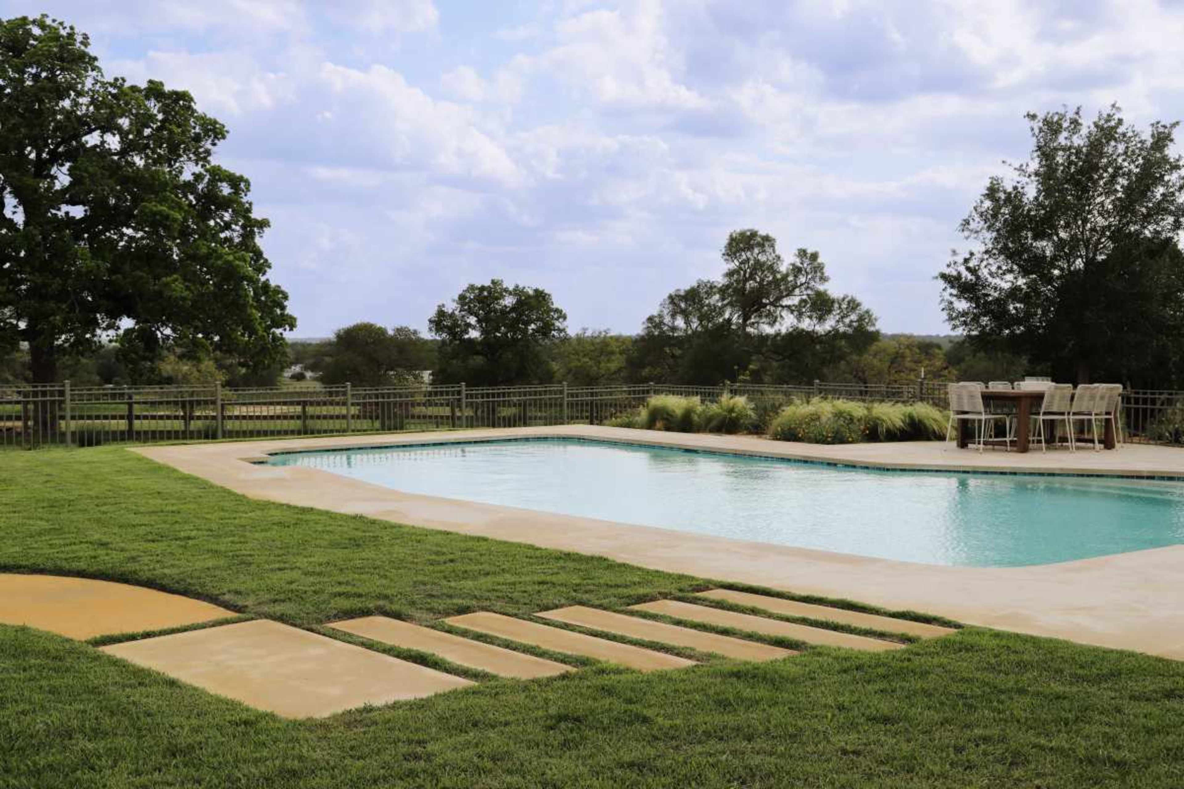A rectangular swimming pool is surrounded by a grassy area and stone pathways, with a dining set visible in the background under a cloudy sky.