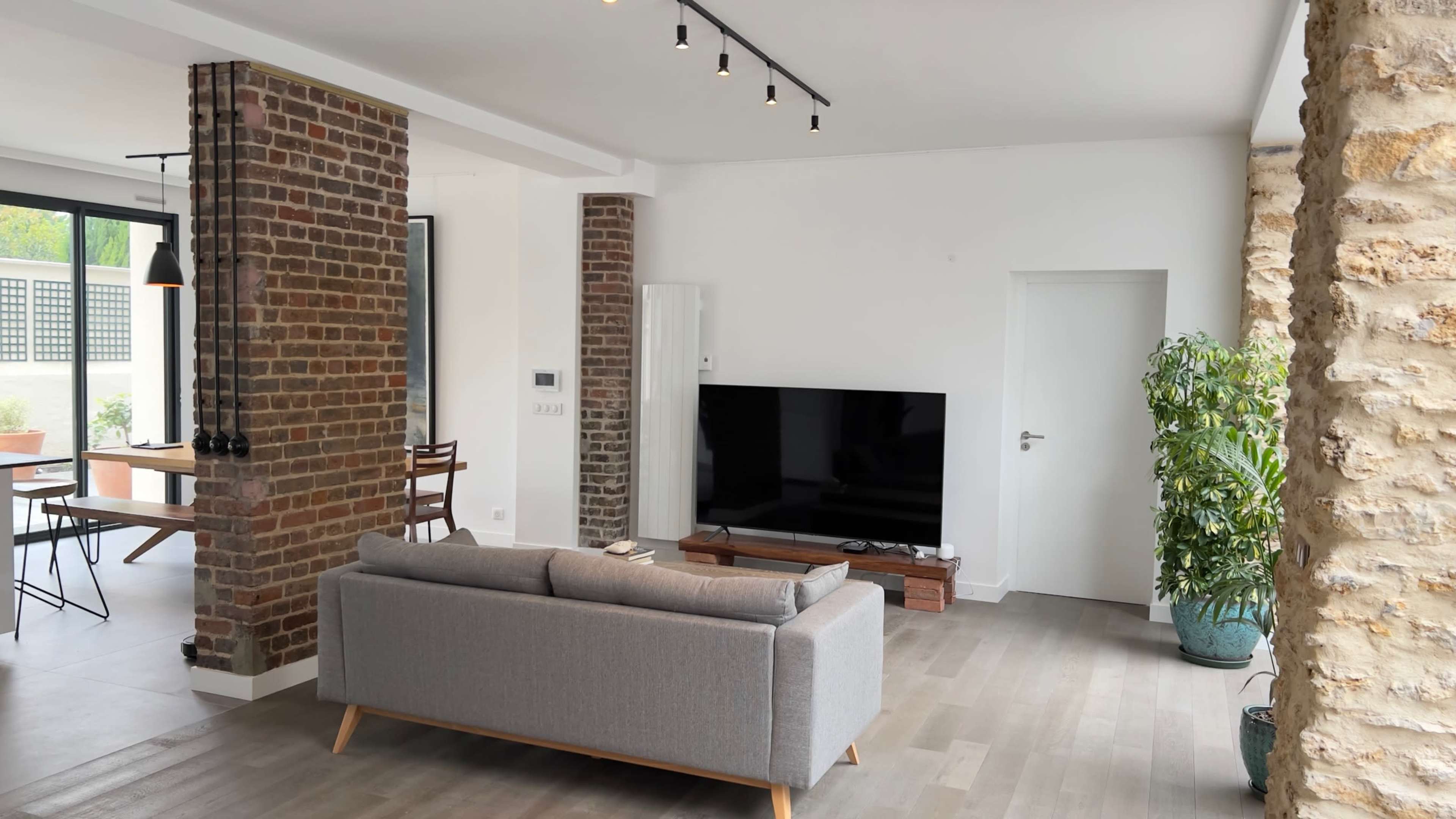 A modern living room with a gray sofa, a flat-screen TV on a wooden stand, exposed brick walls, and a potted plant in the corner.
