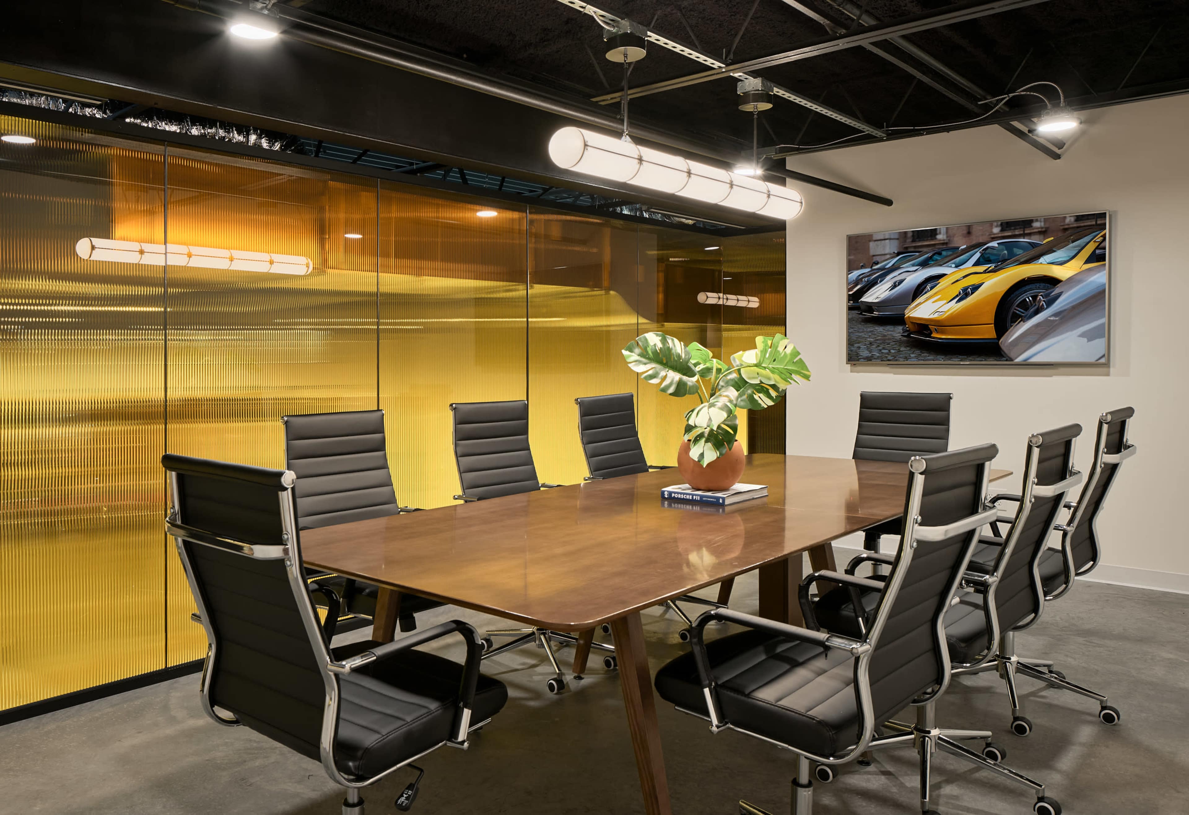 A modern conference room features a large wooden table surrounded by sleek black chairs, with a plant centerpiece and a wall-mounted display showcasing luxury cars.