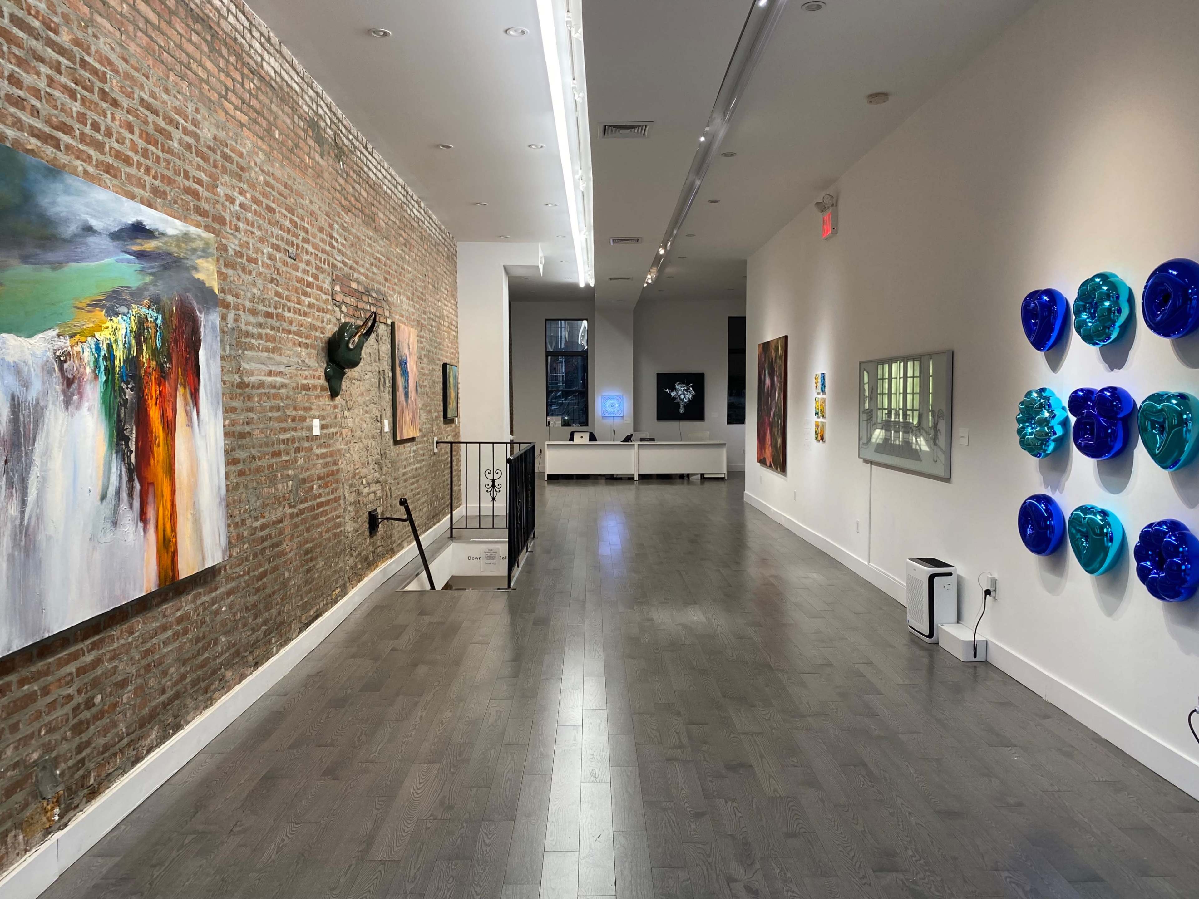 A contemporary art gallery with brick walls featuring various artworks, a staircase, and an area for displaying multimedia pieces.
