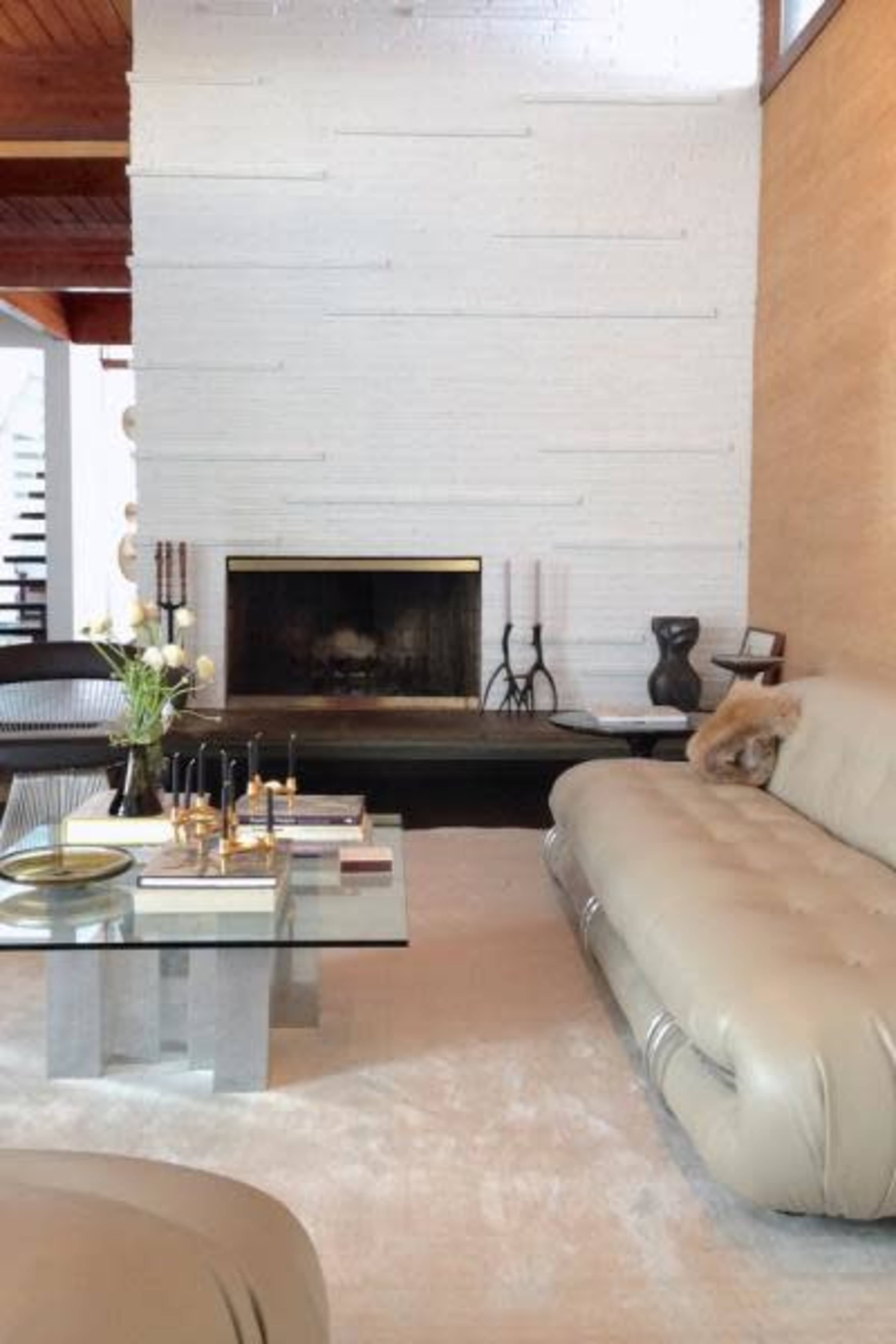 The image shows a modern living room featuring a light-colored sofa, a glass coffee table, and a stone fireplace against a textured wall.