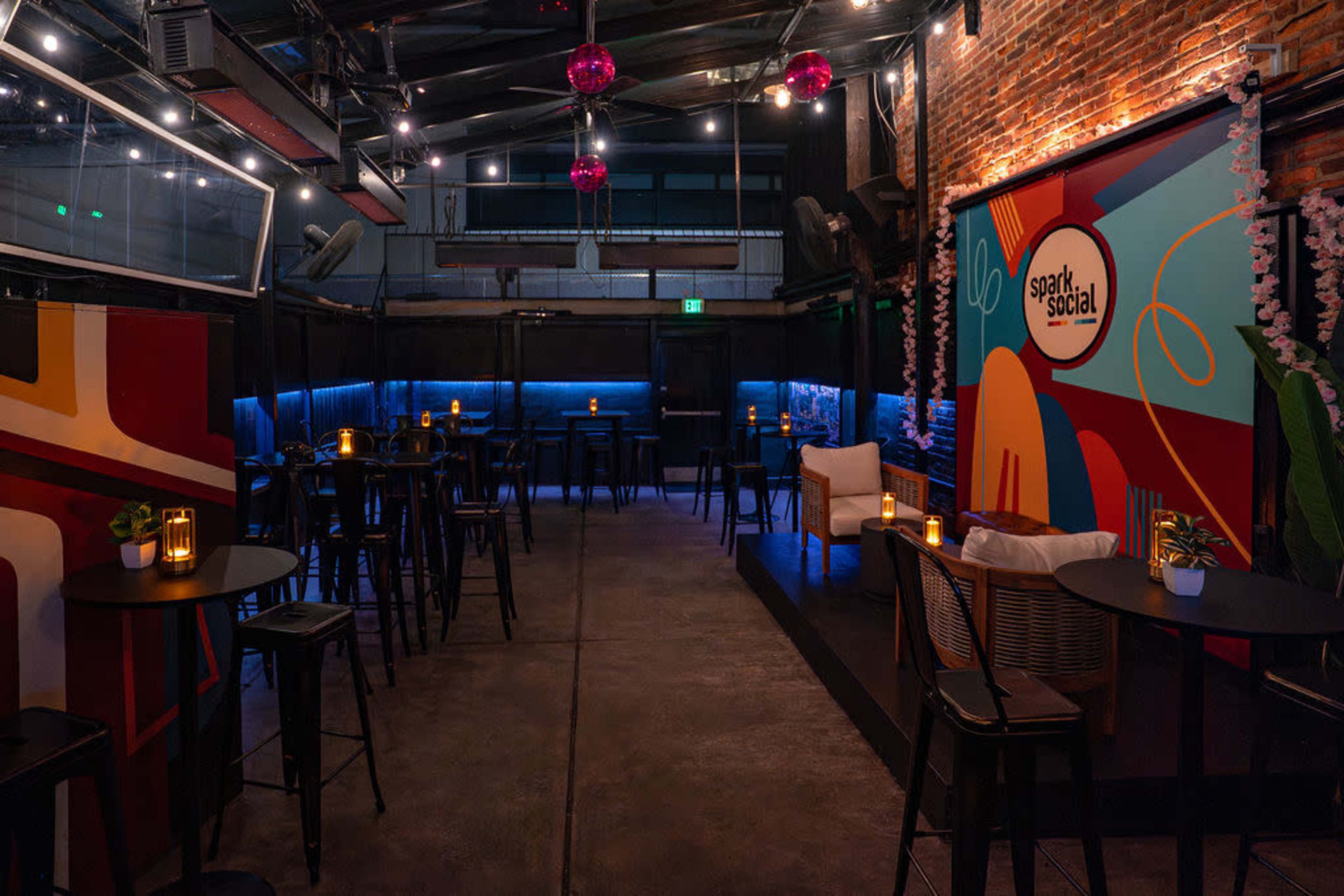 The image shows an indoor venue with a mix of seating arrangements, featuring tables and a couch, illuminated by warm lighting and decorated with colorful murals and hanging fixtures.