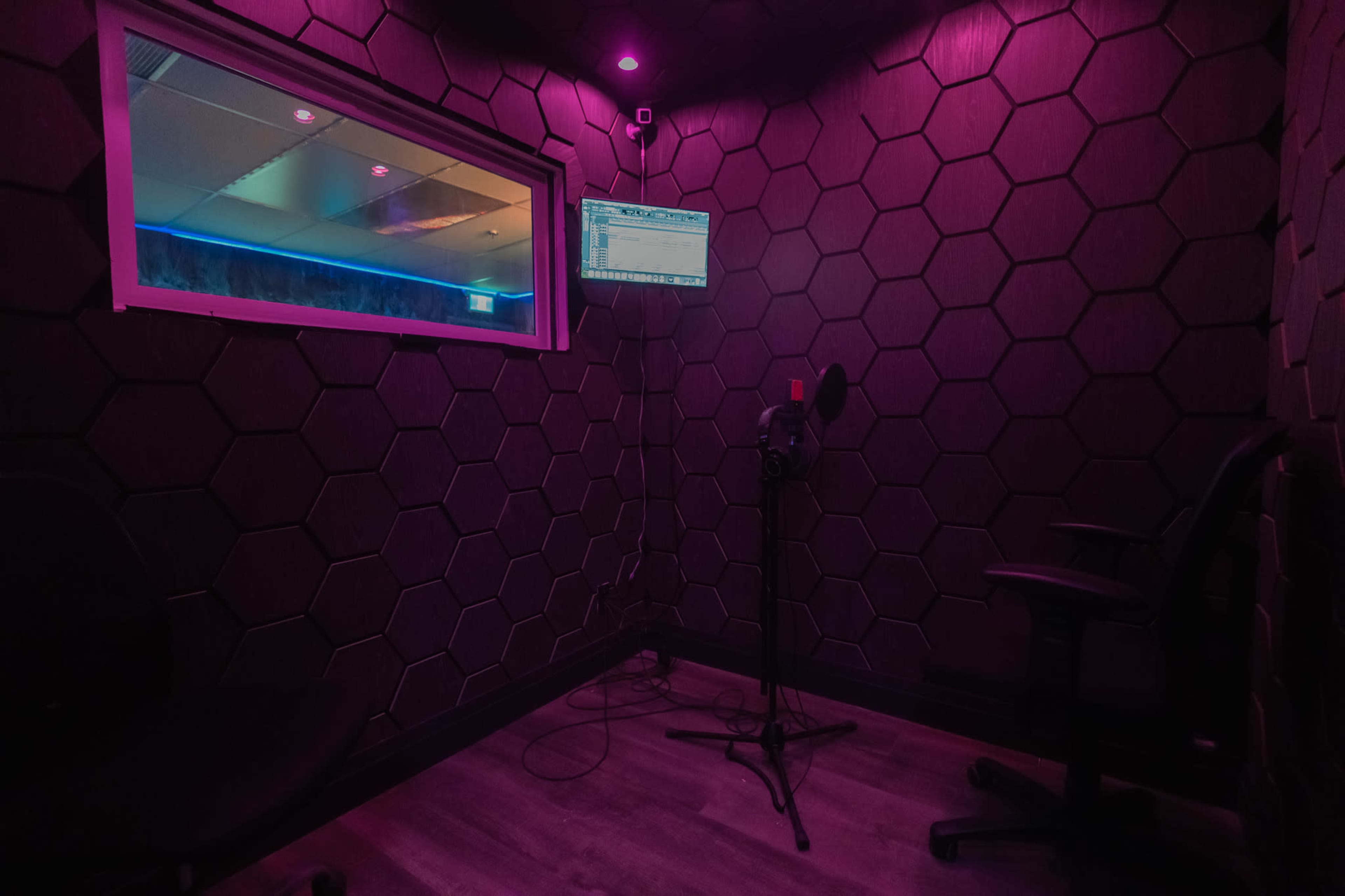 The image shows a small recording studio booth with hexagonal patterned walls, a microphone on a stand, and a monitor displaying audio software.