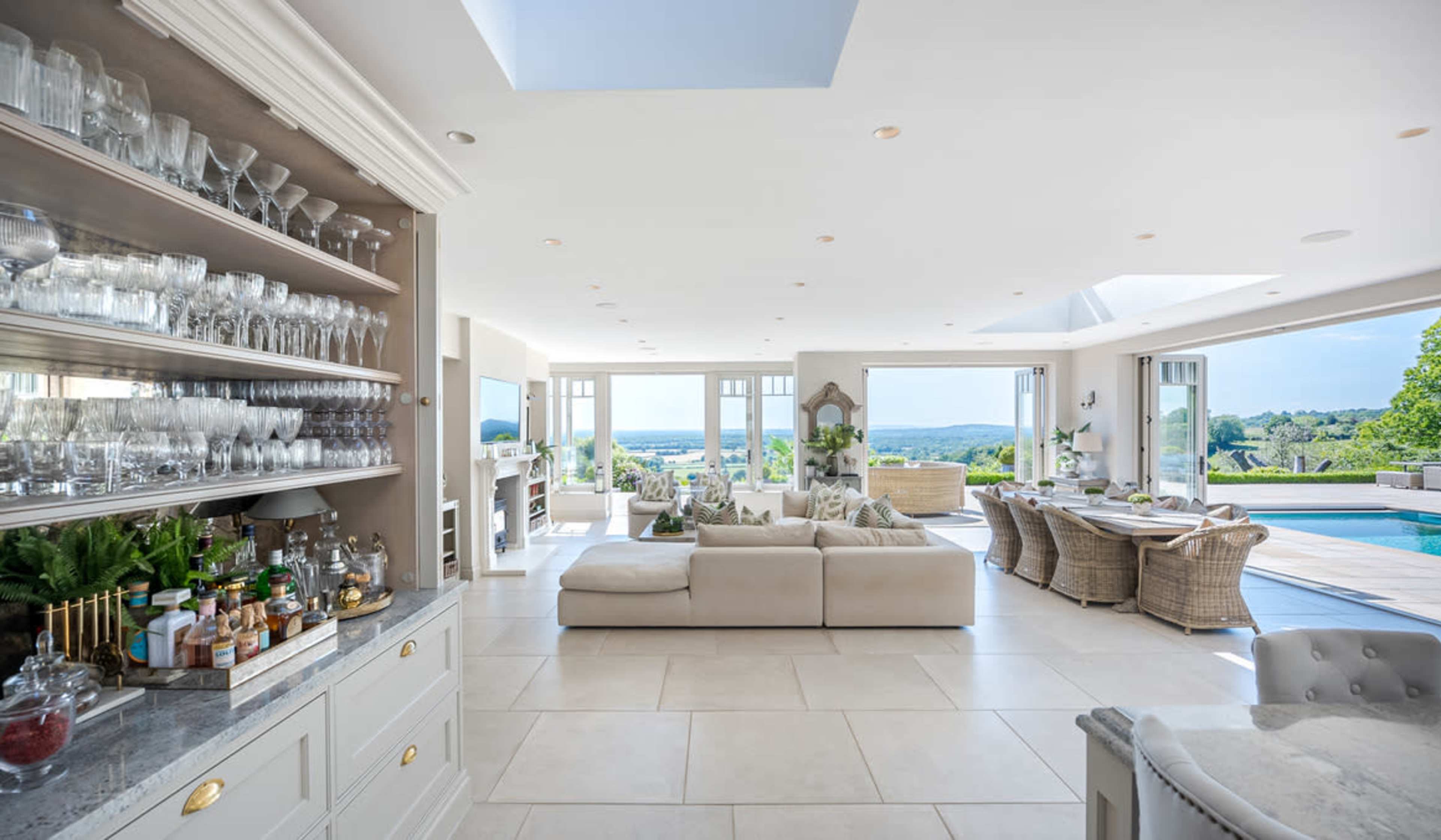 The image shows a spacious modern living area with a large sectional sofa, a glassware display, and expansive windows offering a view of the outdoors.