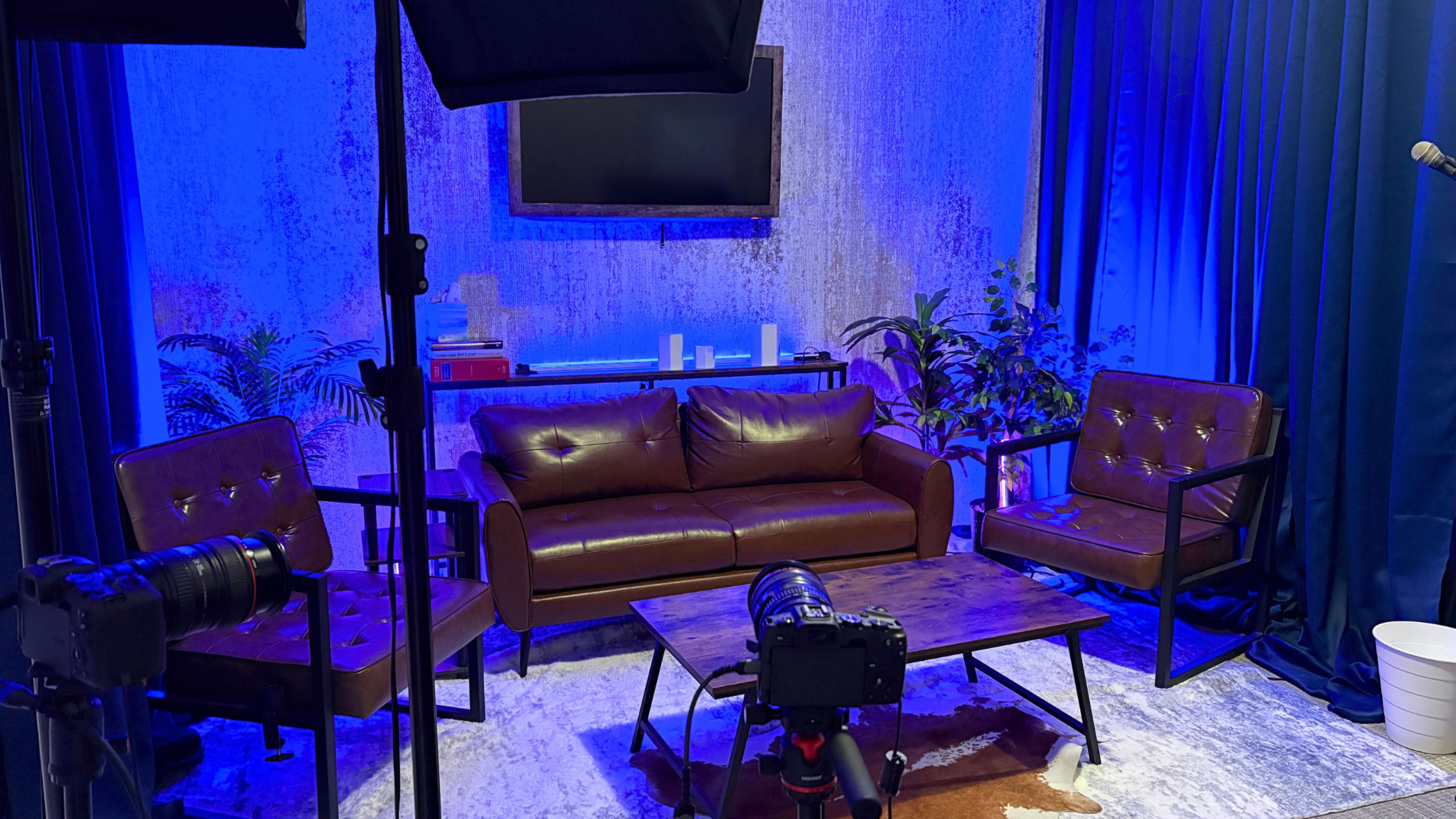 A well-lit studio setup features a leather sofa, two chairs, a coffee table, and a television, surrounded by plants and blue lighting.