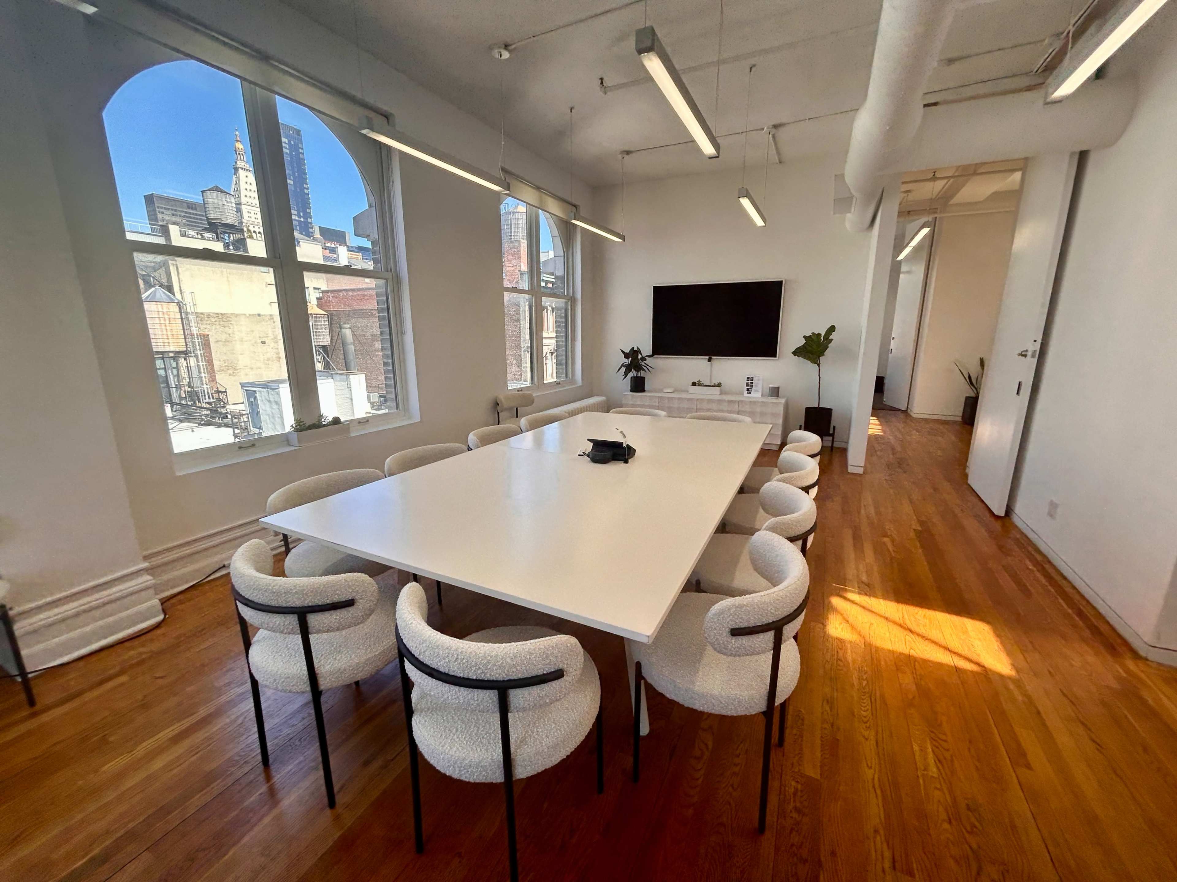 A bright conference room features a large white table surrounded by upholstered chairs, with large windows offering a view of the city skyline.