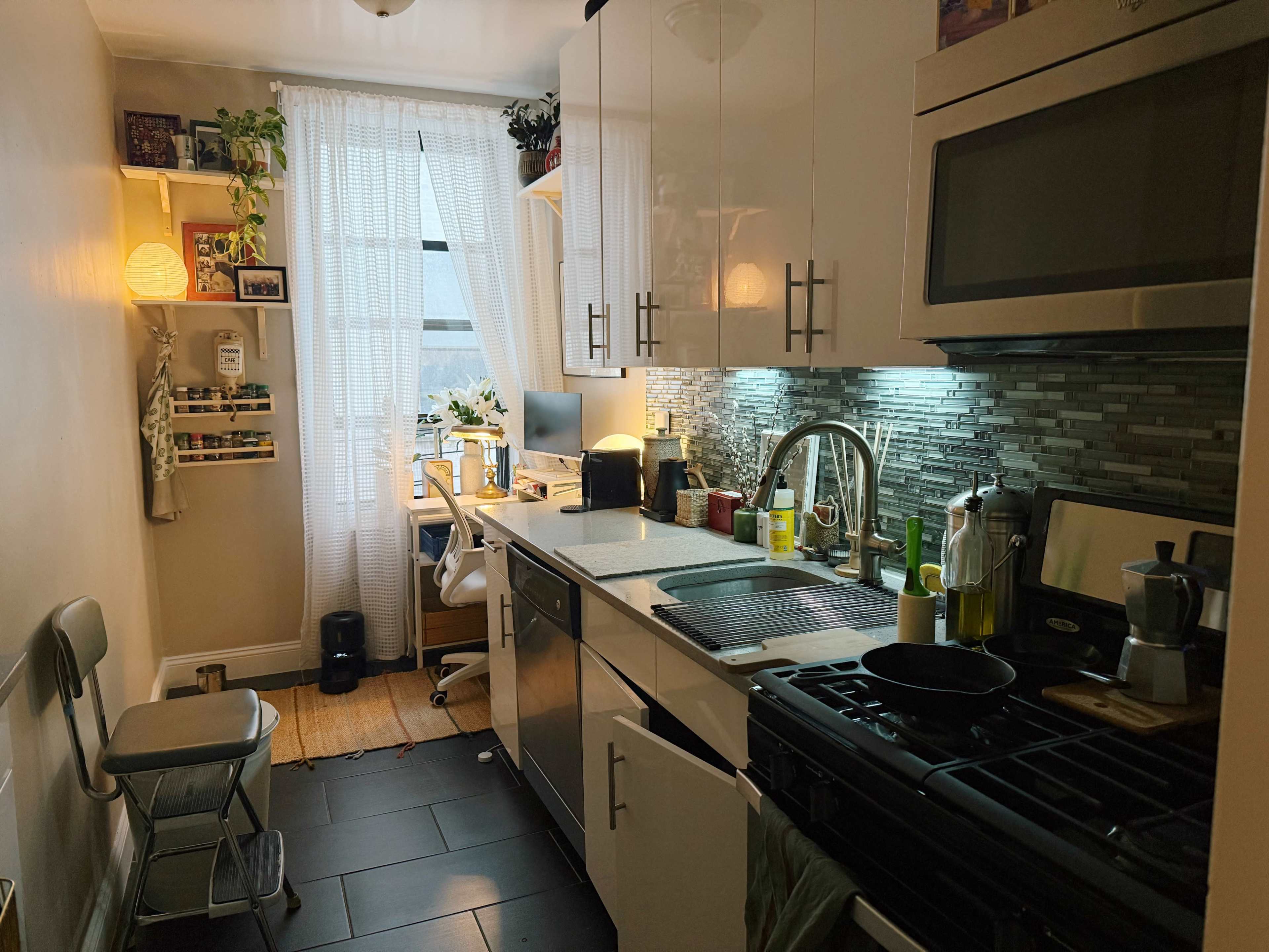 The image shows a bright kitchen with modern cabinets, a sink, a stovetop, and a small dining area, featuring a window with sheer curtains.