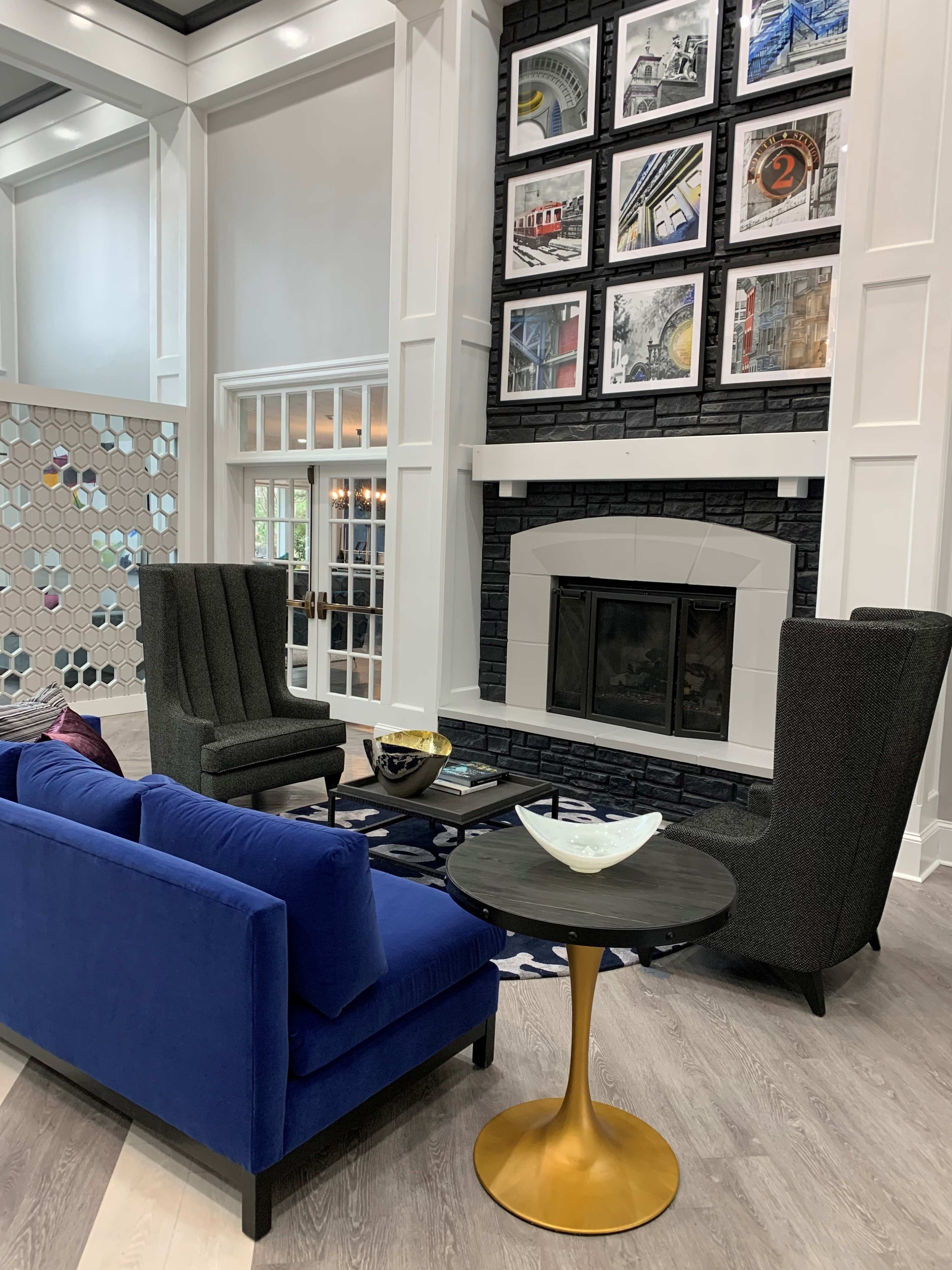 The image shows a modern living area with a blue sofa, two high-backed chairs, a round table, and a wall adorned with framed artwork above a black stone fireplace.