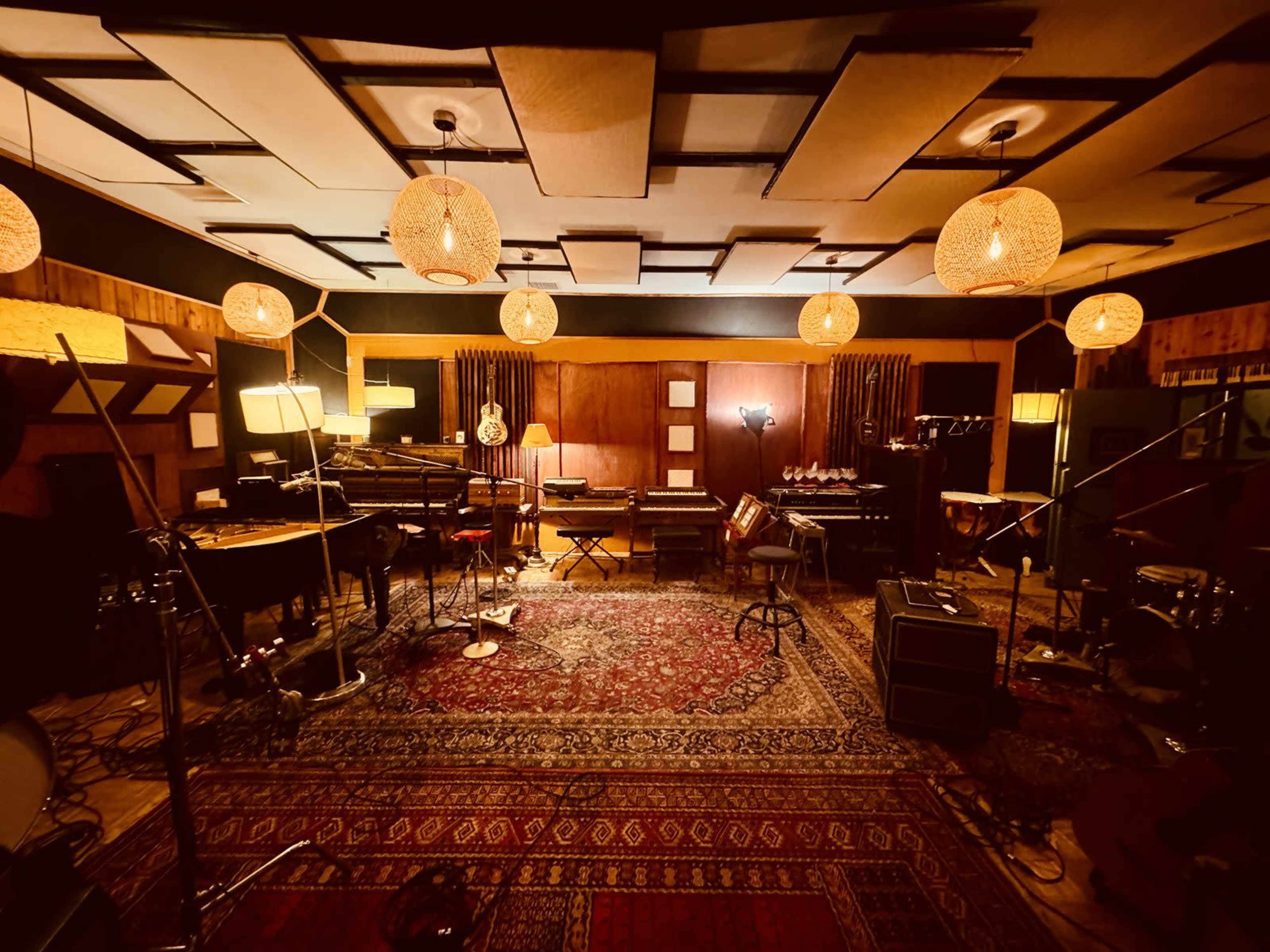 The image shows a well-lit music recording studio featuring an array of musical instruments, sound equipment, and various lighting fixtures.