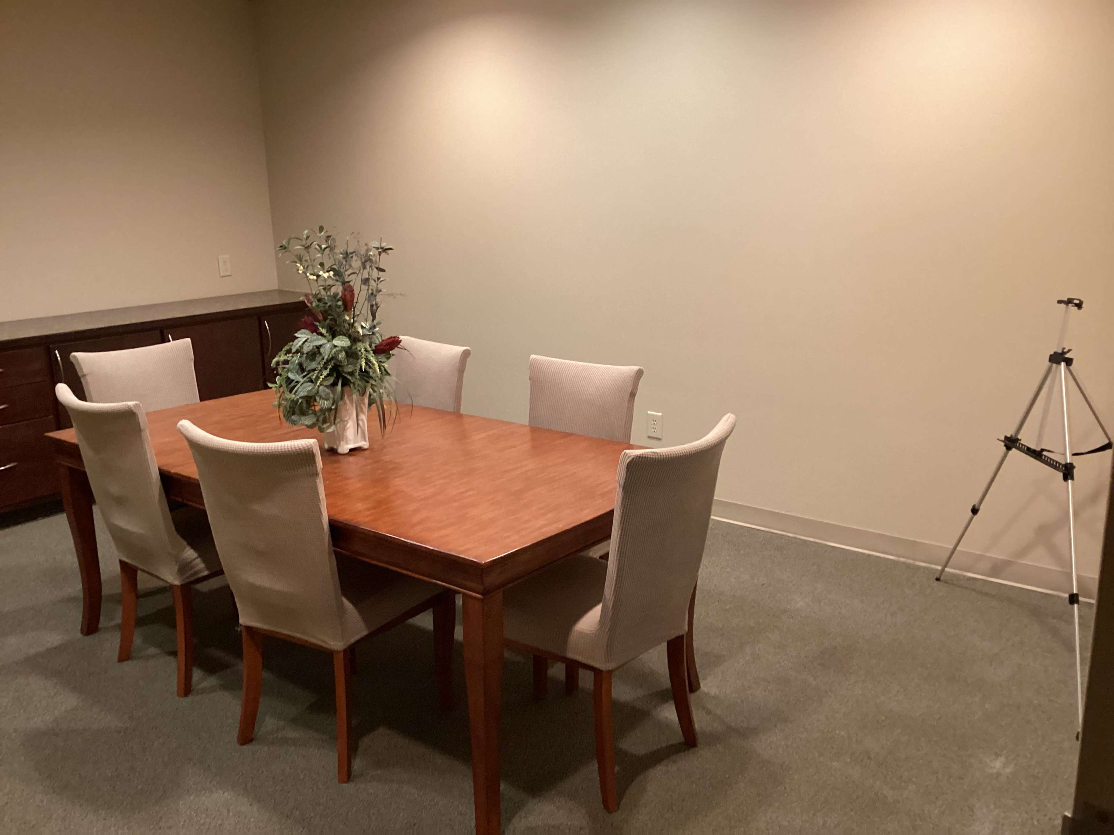 A wooden dining table surrounded by six chairs is set in a sparsely furnished room, with a potted plant at the center and a tripod in the corner.