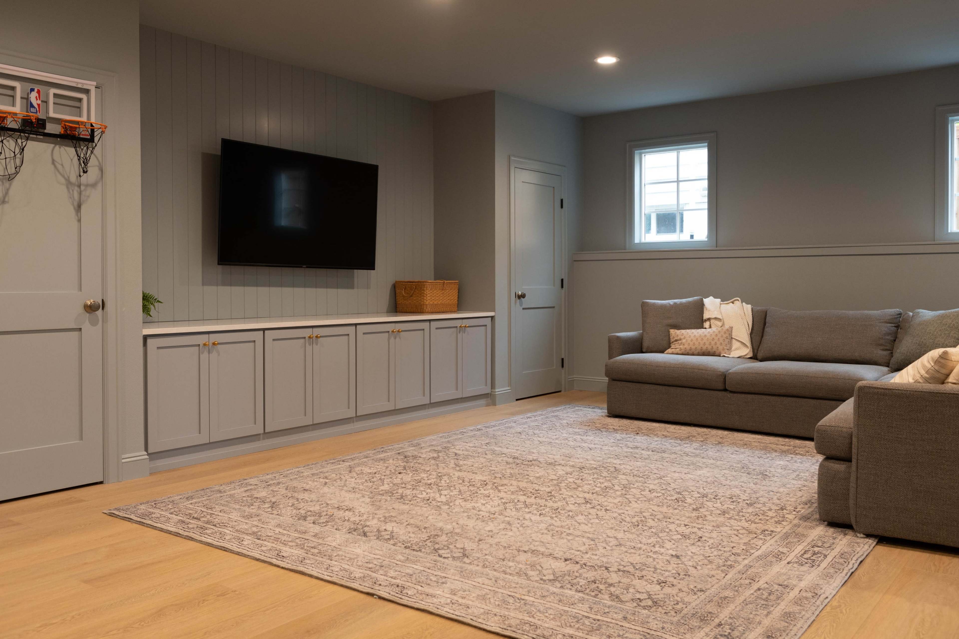 A modern, well-lit living room features a gray sectional sofa, a decorative area rug, a media console beneath a wall-mounted TV, and two windows allowing natural light.