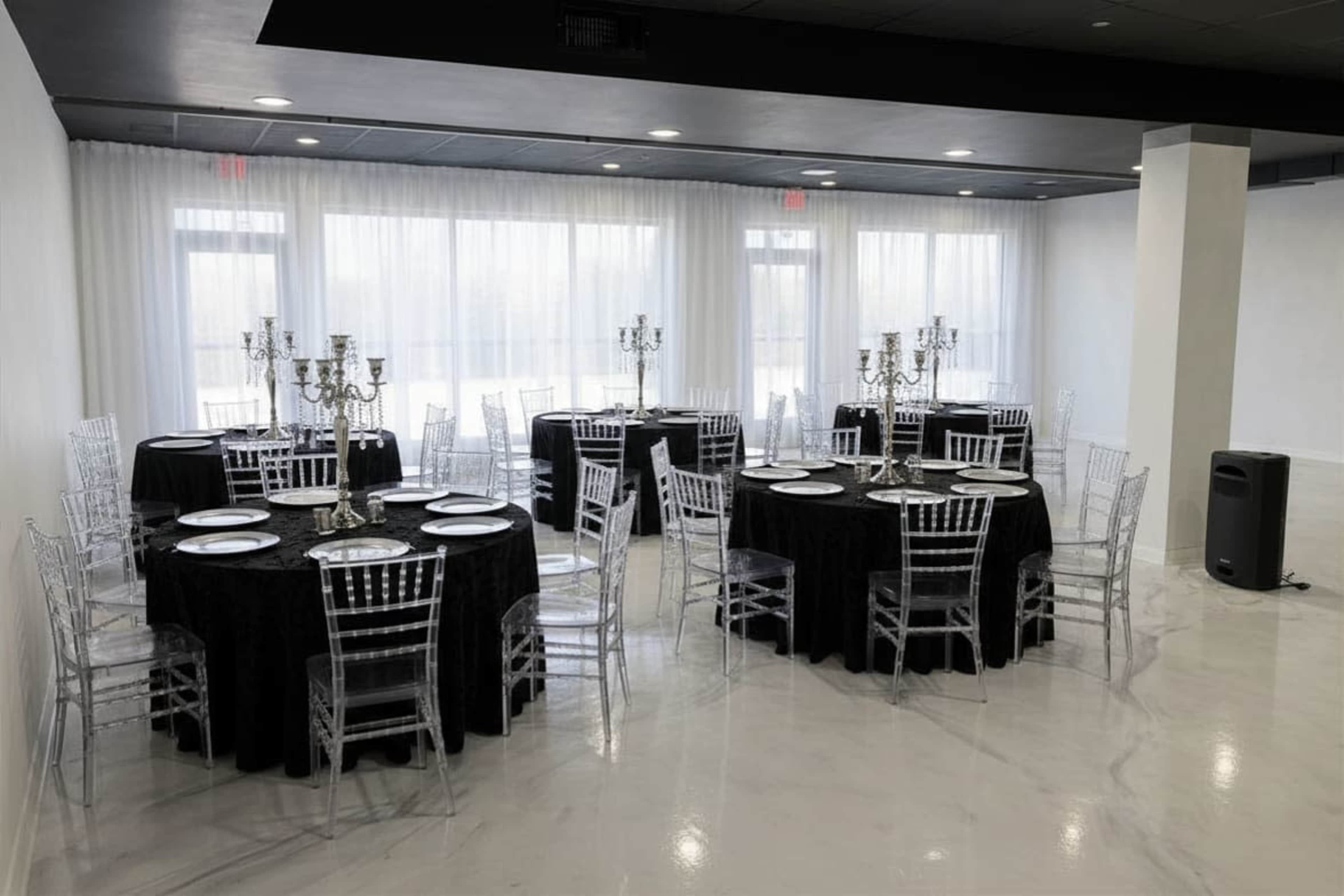 The image shows a spacious event room with several round tables covered in black tablecloths and decorated with elegant candle holders, set against large, bright windows.