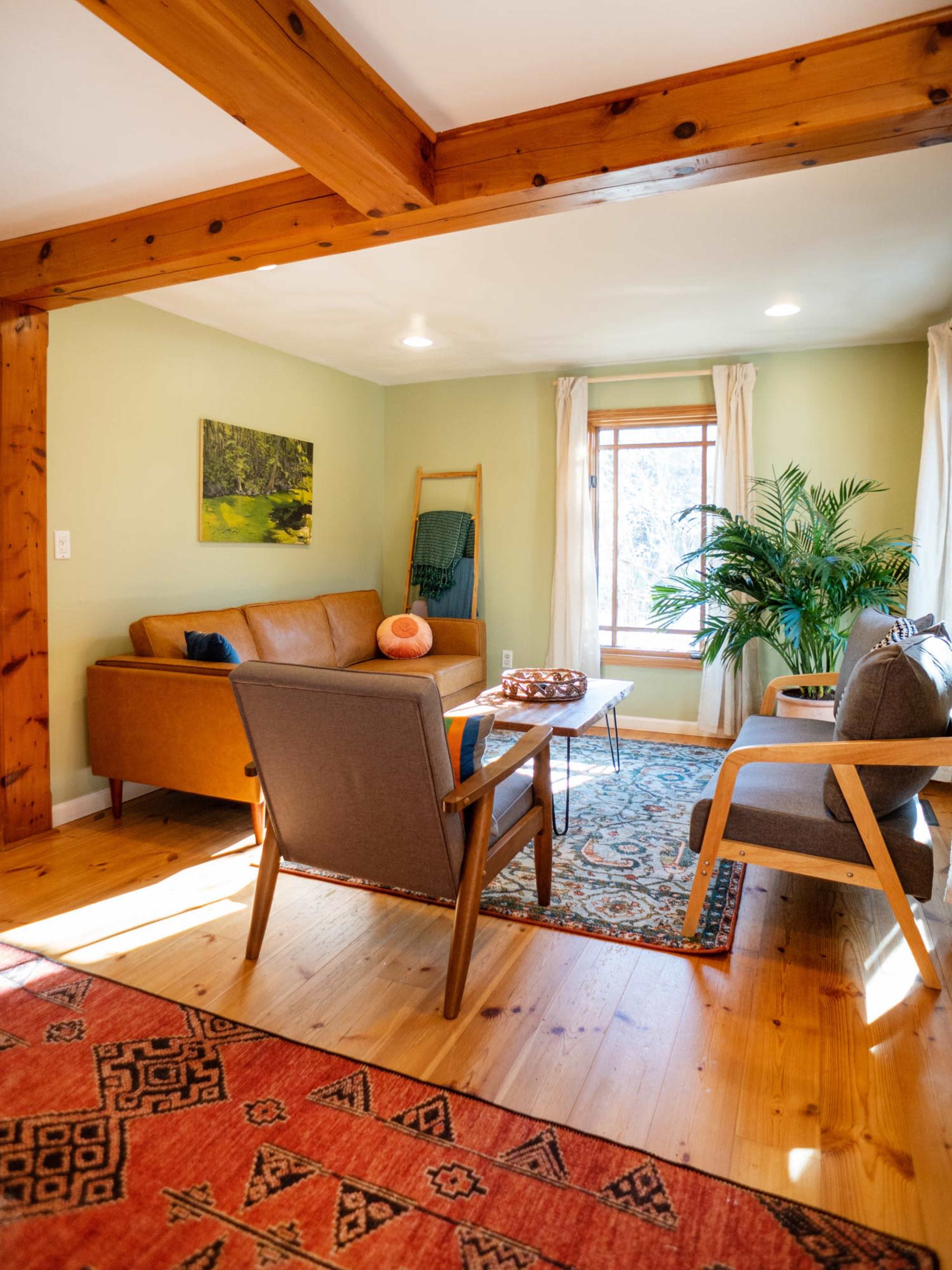 A cozy living room features a brown leather couch, armchairs, a wooden coffee table, and a large window with natural light illuminating the space.